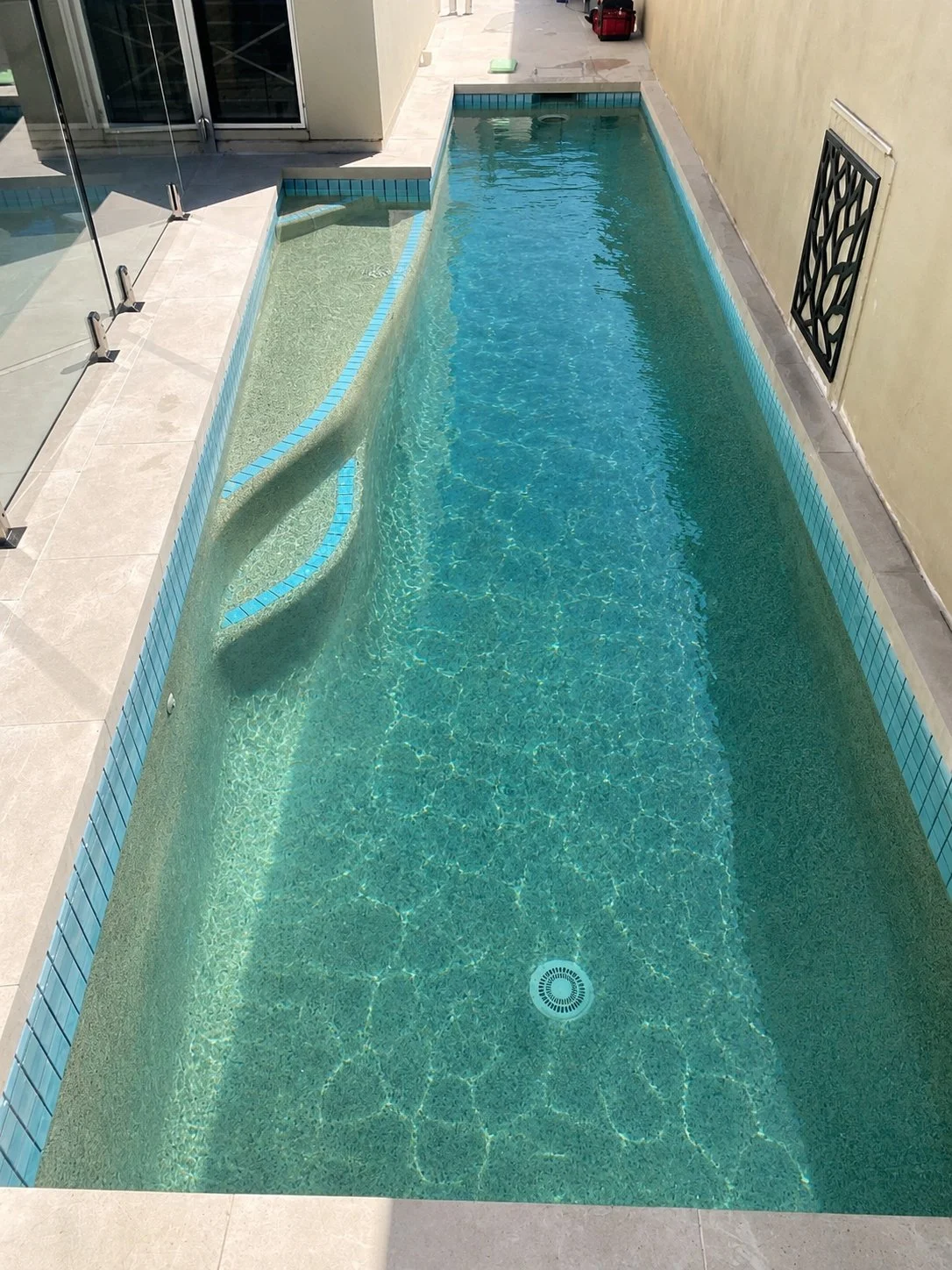 Narrow lap pool with curved entry steps, blue waterline tiles and glass fencing beside a house in Parrearra.