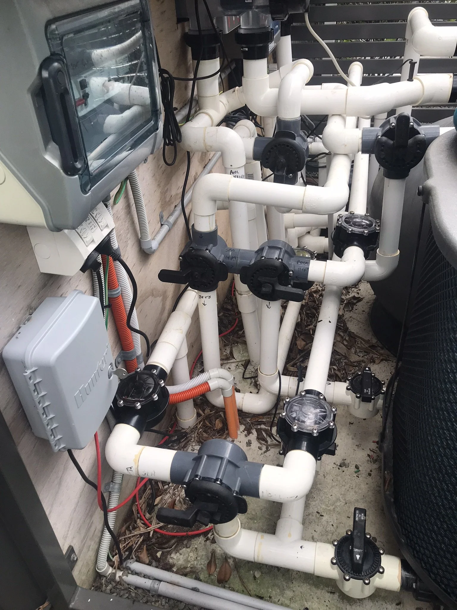 Dense network of white PVC pool pipes, elbows and black valves beside electrical control boxes.