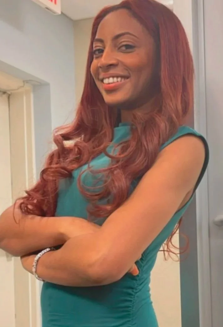 Gabriella St. Fleur, the owner and founder, with long, wavy red hair, smiling, wearing a teal sleeveless dress, standing with arms crossed inside a building.
