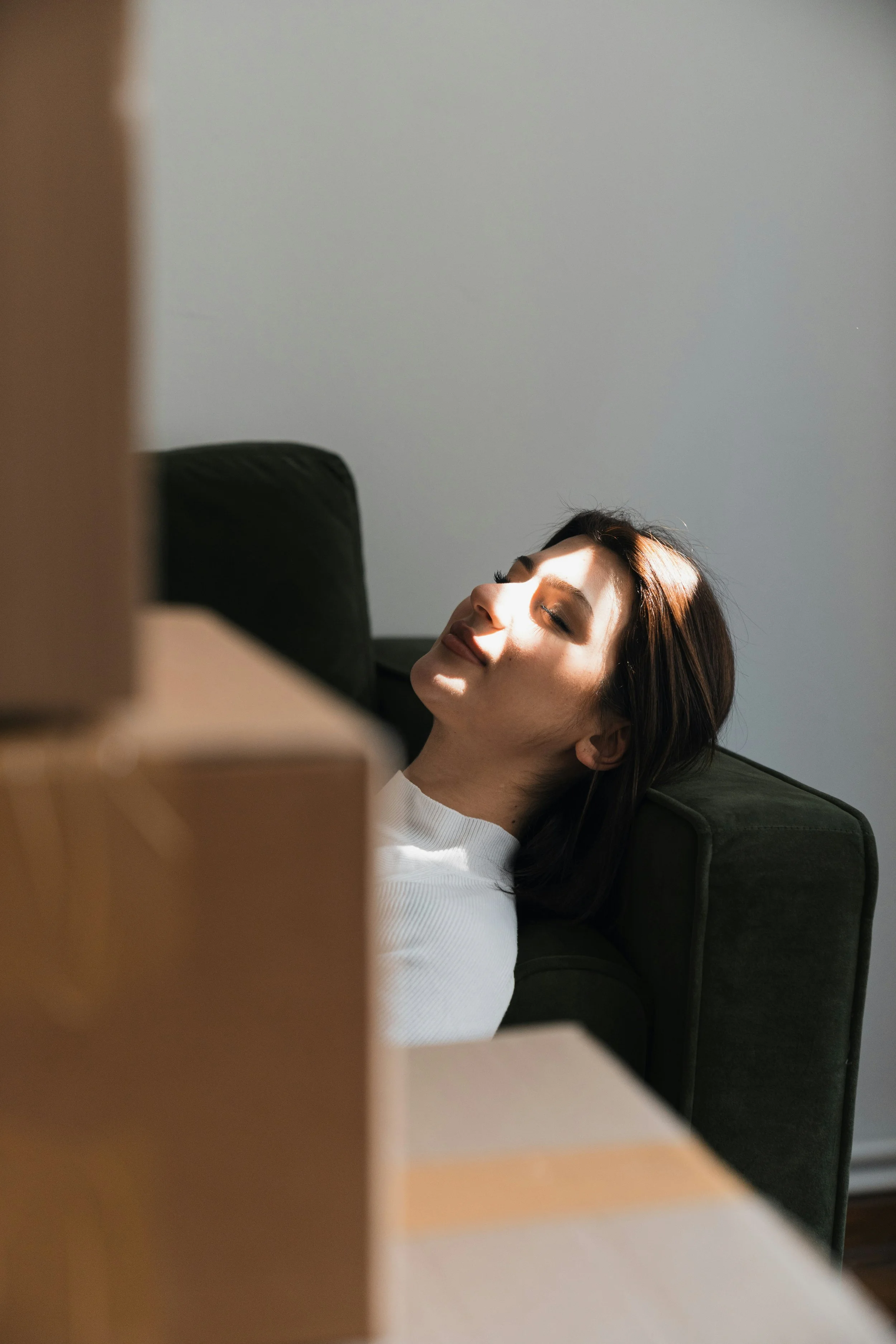A woman with short brown hair relaxing on a dark green sofa with her eyes closed, bathed in natural sunlight.