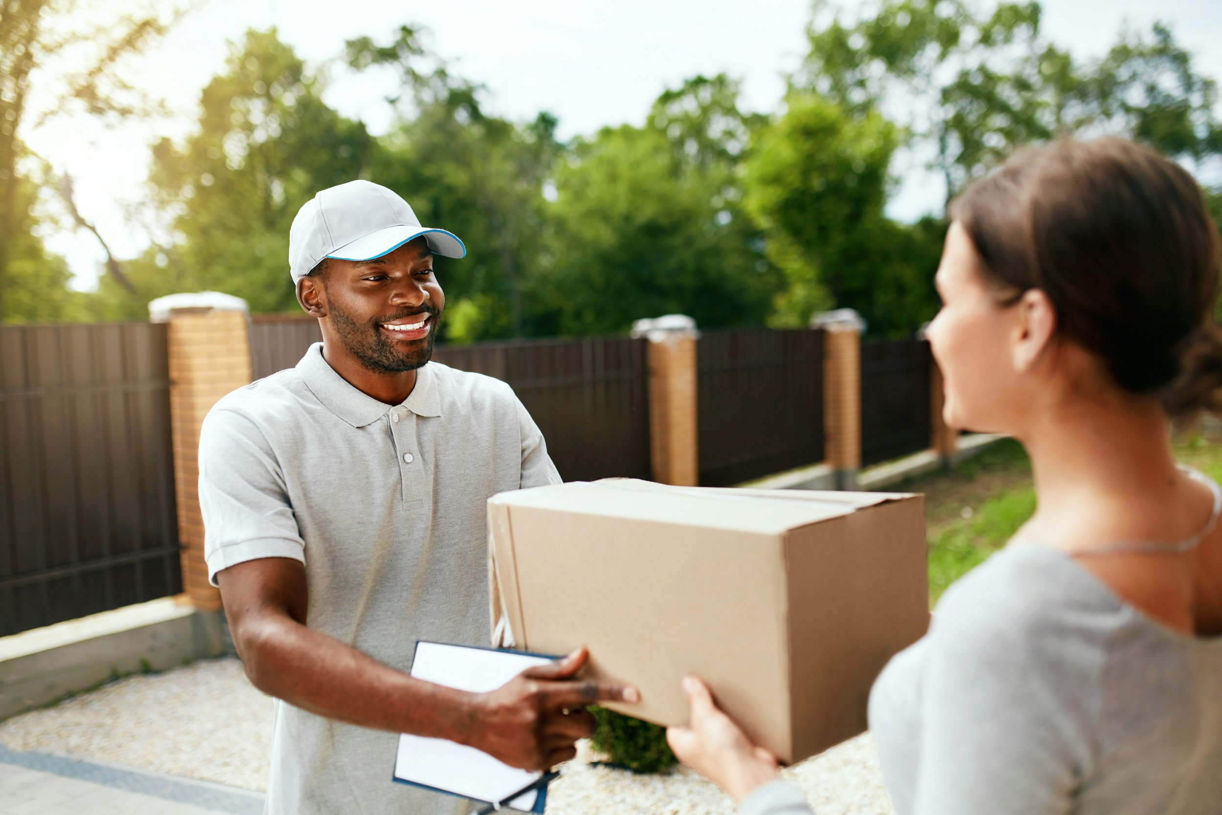 A man in a gray shirt and cap is smiling as he receives a cardboard box from a woman with brown hair, both standing outdoors in front of a black fence with brick posts and green trees.