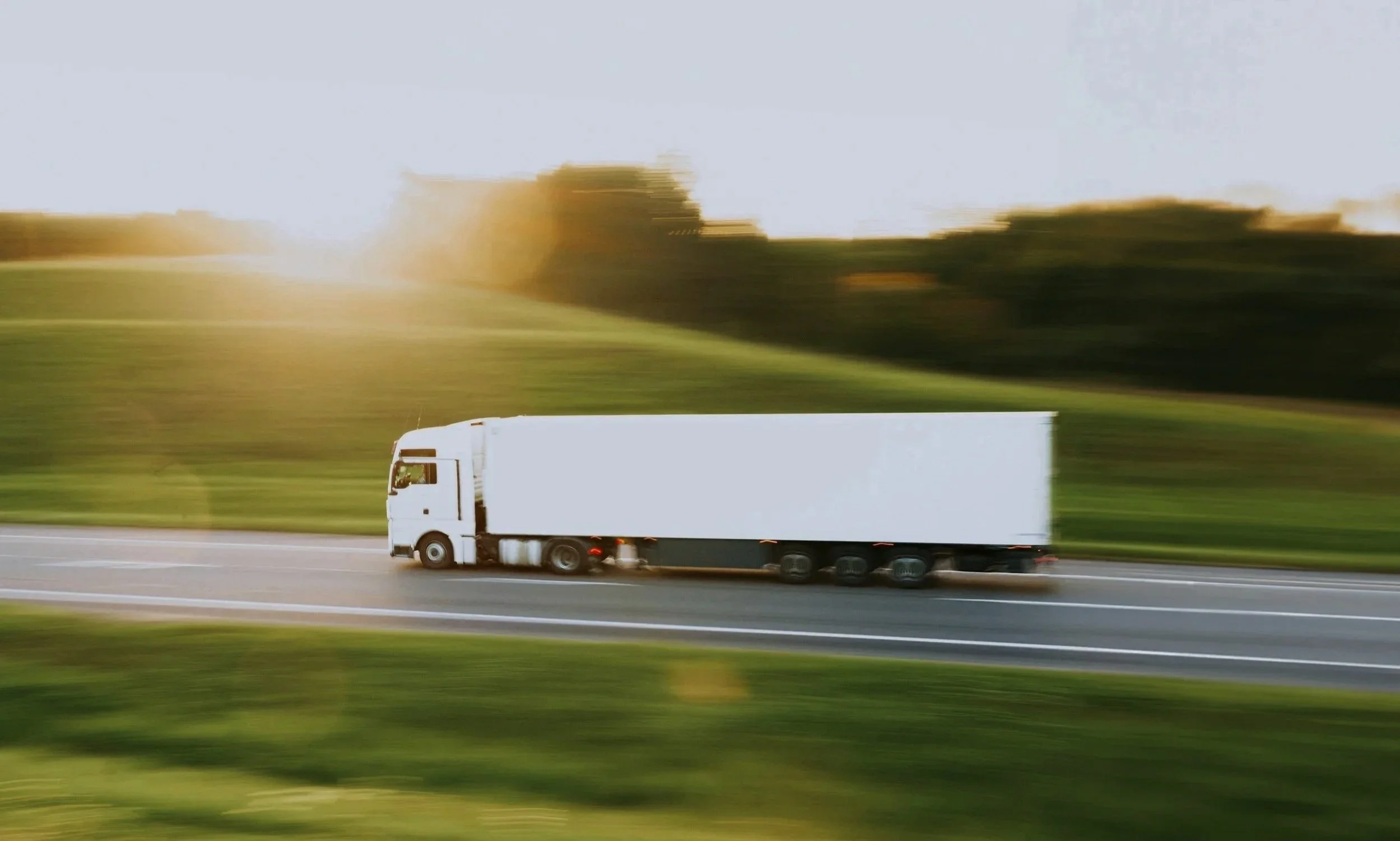A semi-truck driving on a highway with mountainous terrain and cloudy sky in the background.