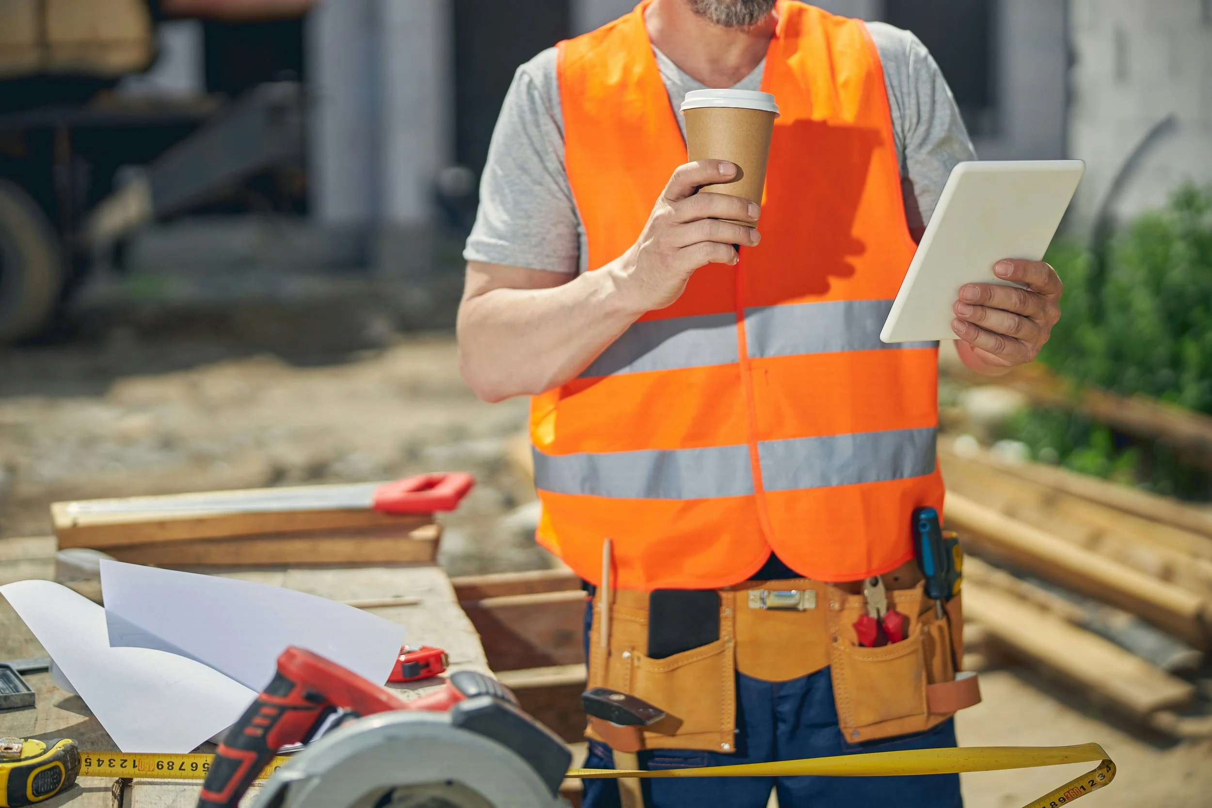 Construction worker wearing an orange safety vest holding a coffee cup and a tablet at a construction site.