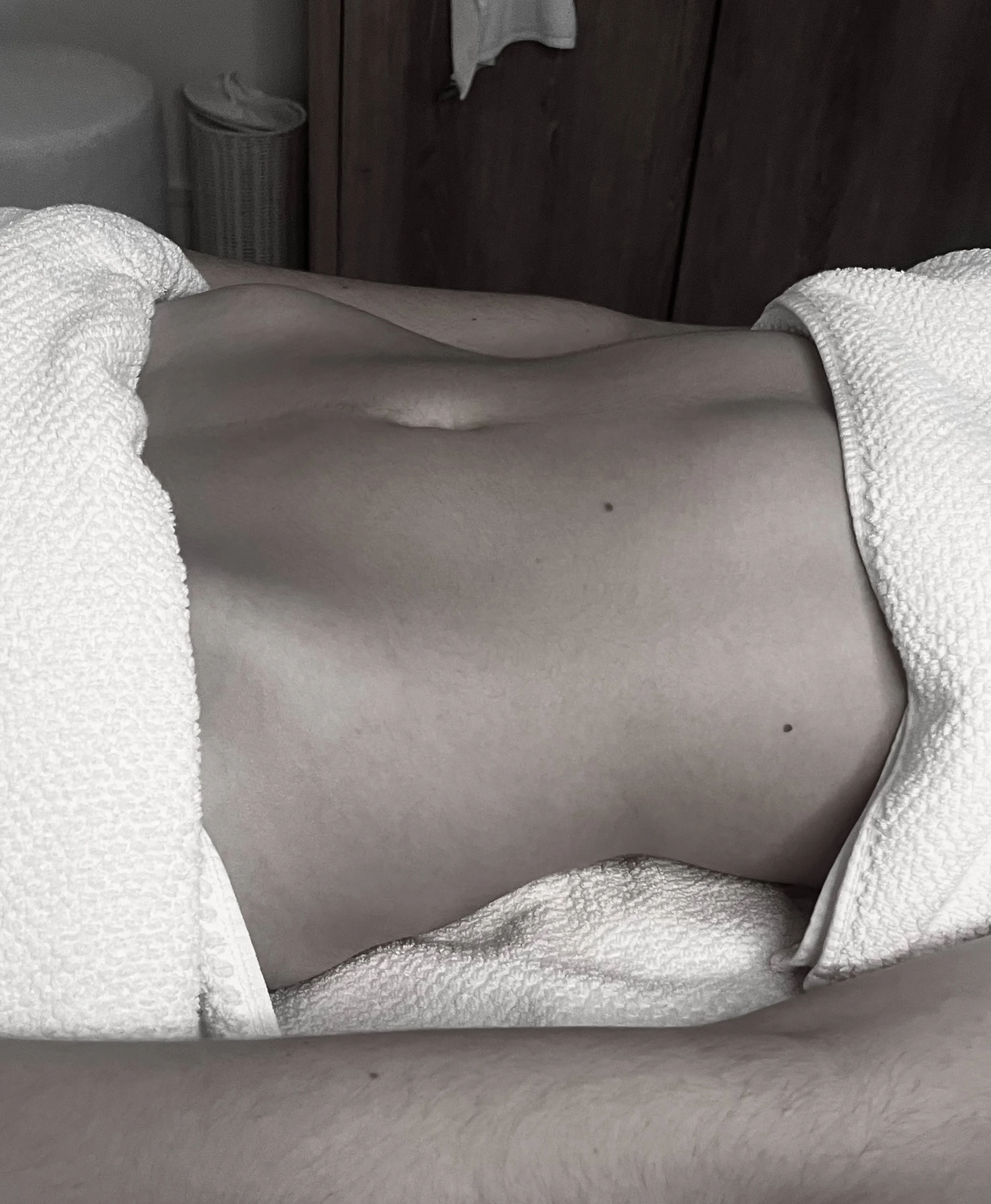 A person's midsection lying on a white textured towel or blanket, with a wooden wardrobe and other household items in the background.