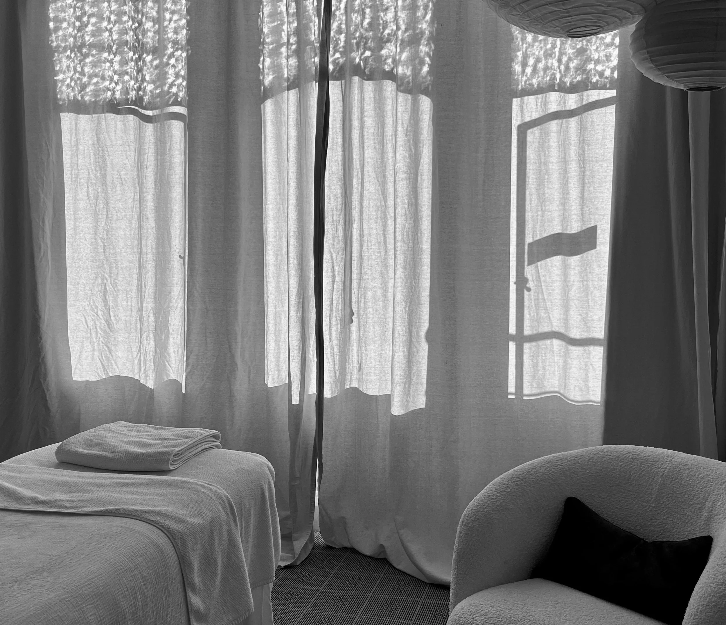 A black-and-white photo of an interior room with a bed, a towel, a chair with a cushion, and an open window with curtains and blinds casting shadows.