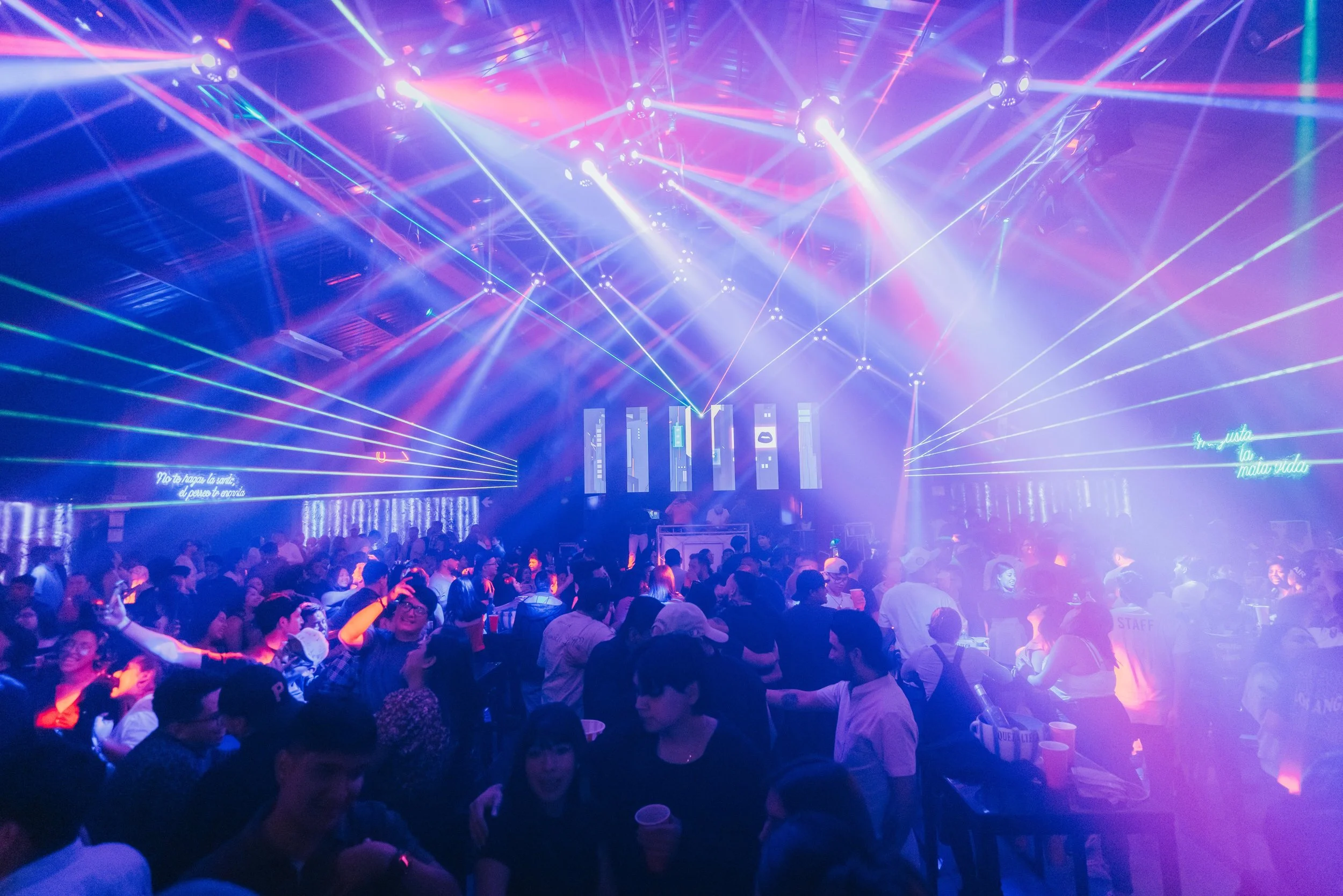 A crowded nightclub with colorful laser lights and a stage with vertical screens displaying graphics.