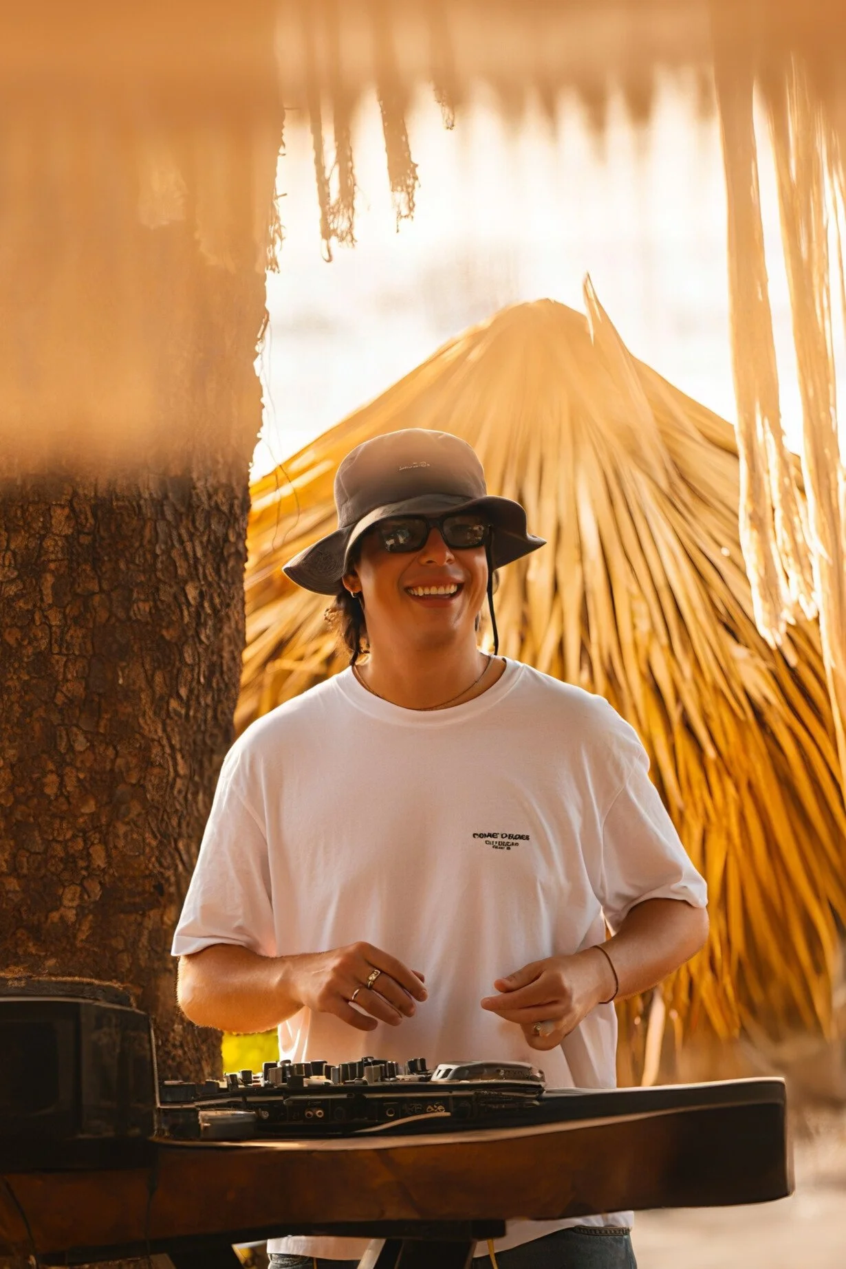 A woman smiling, wearing sunglasses, a bucket hat, and a white t-shirt, standing behind a DJ console with a thatched roof and palm trees in the background.