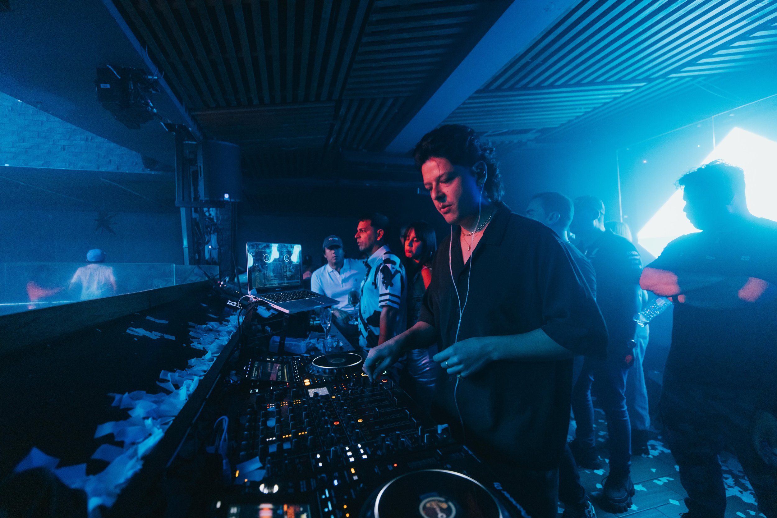 A DJ performs in a nightclub with blue lighting, surrounded by people dancing and socializing.