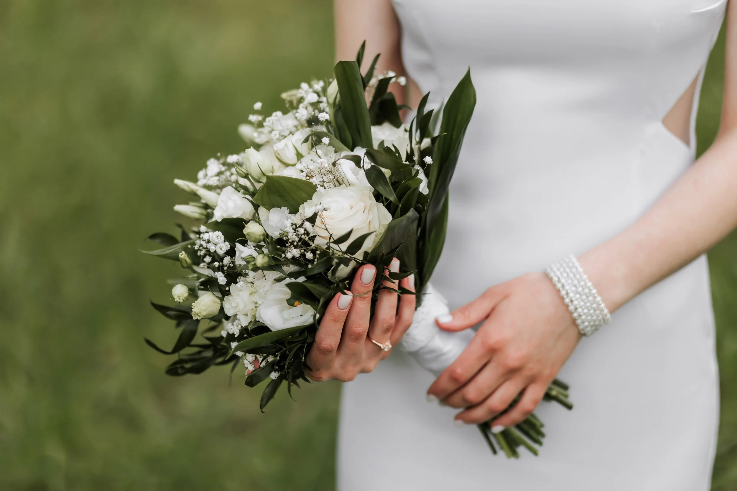 A bride in a white dress holding a bouquet of white flowers and greenery, with a pearl bracelet on her wrist.