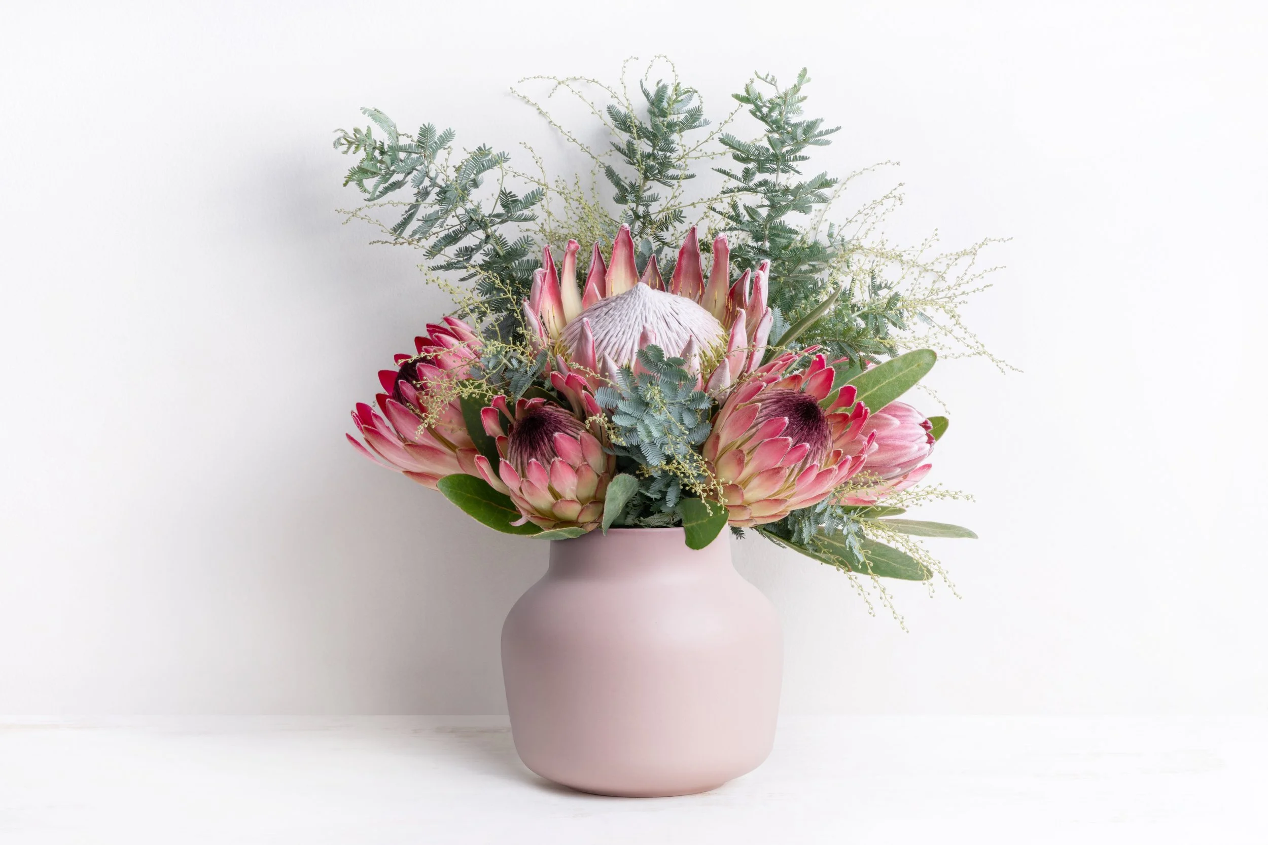 A pink vase with a bouquet of pink protea flowers and green foliage against a white background.