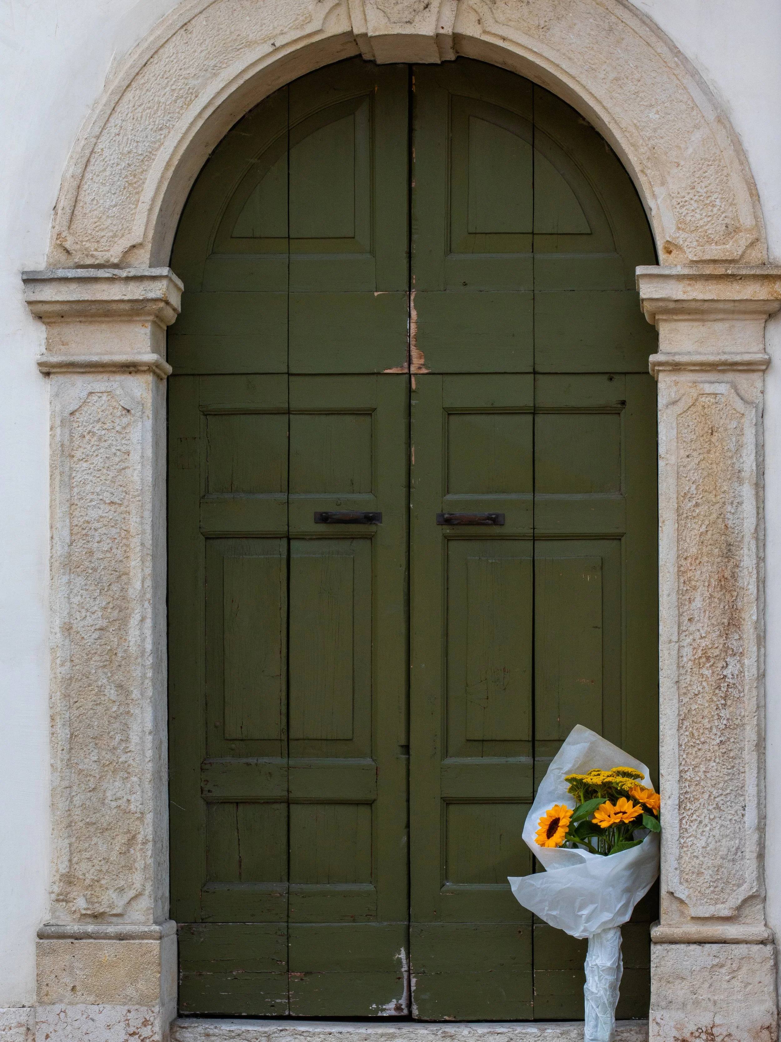 A set of green wooden double doors with an arched stone frame, a bouquet of yellow sunflowers wrapped in white paper leaning against the right side of the door.
