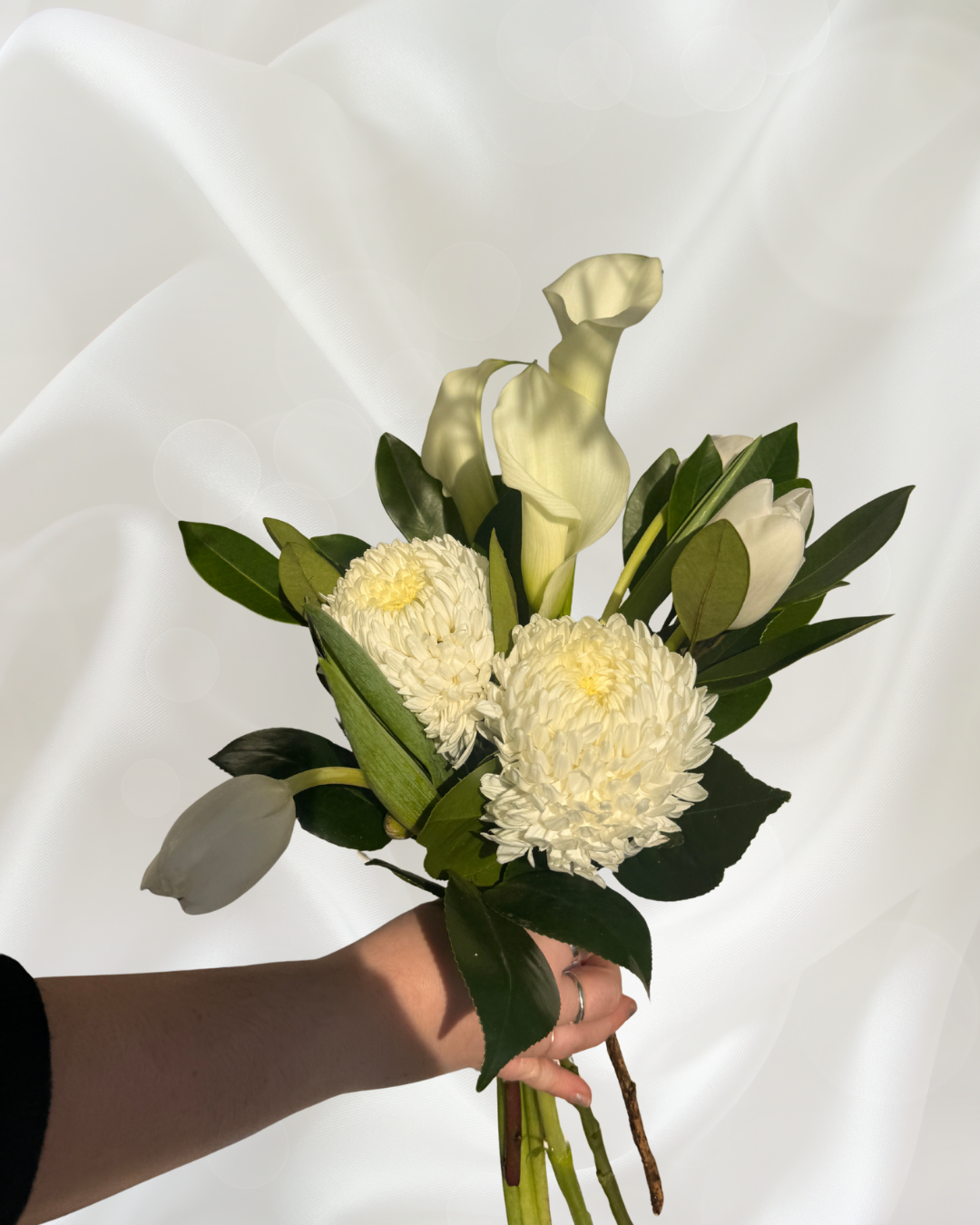 A bouquet of white flowers including calla lilies, tulips, and chrysanthemums, held in a person's hand against a light background with soft lens flare.