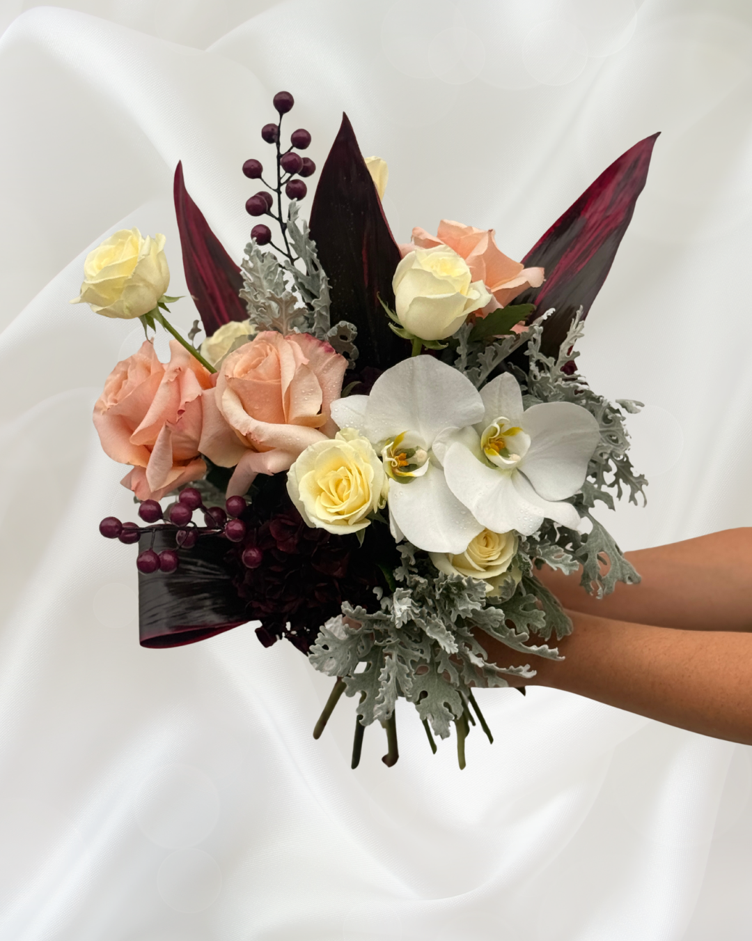 A bouquet of mixed flowers including white orchids, peach roses, white roses, dark red leaves, purple berries, and silver-gray foliage, held in a person's hand with a white background.