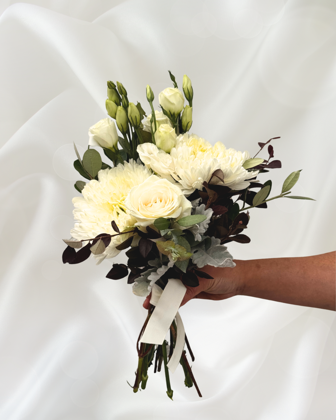 A hand holding a bouquet of white roses, white dahlias, and greenery against a white satin background.