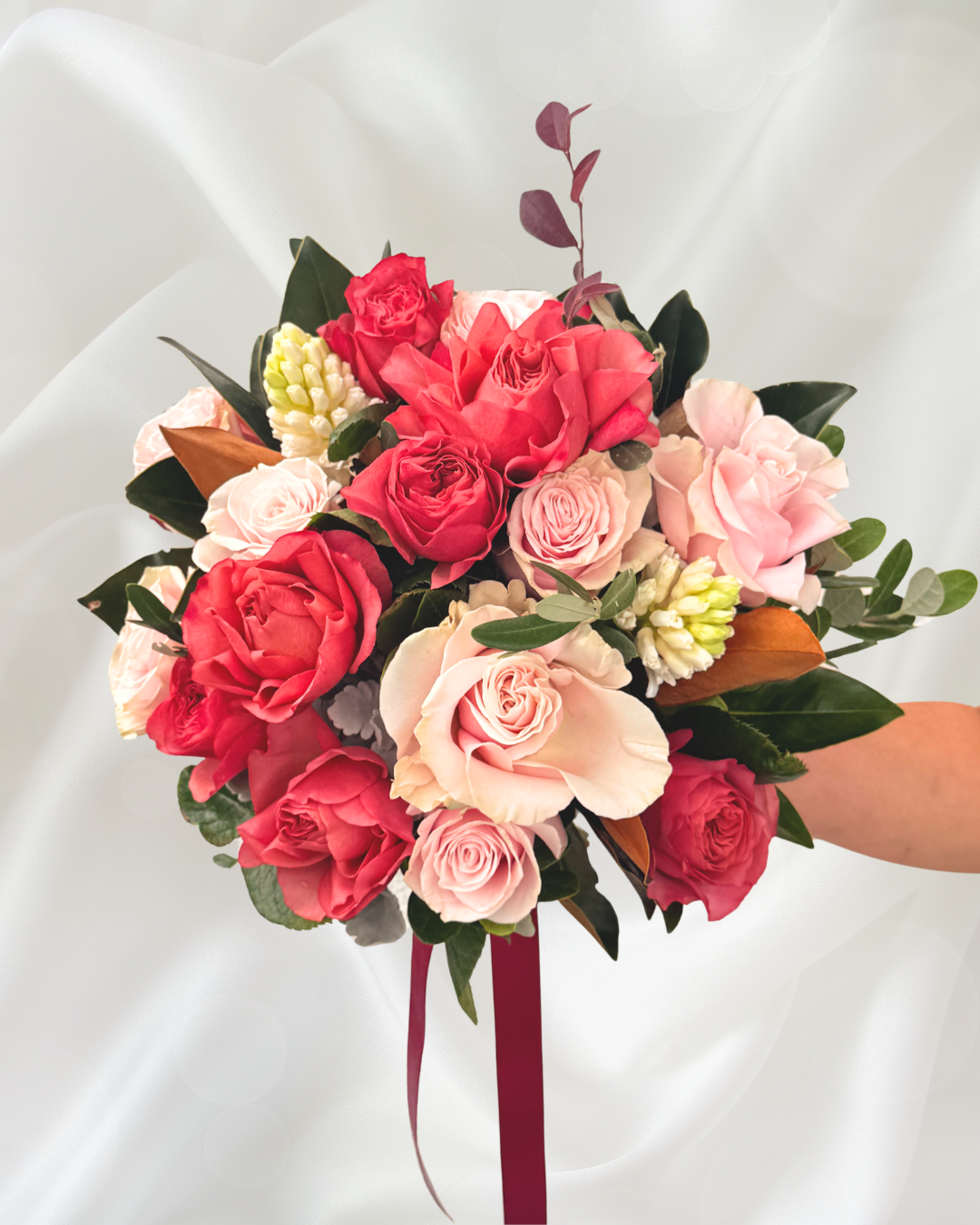 A bouquet of pink, red, and cream roses with greenery, held in a person's hand against a white satin background.