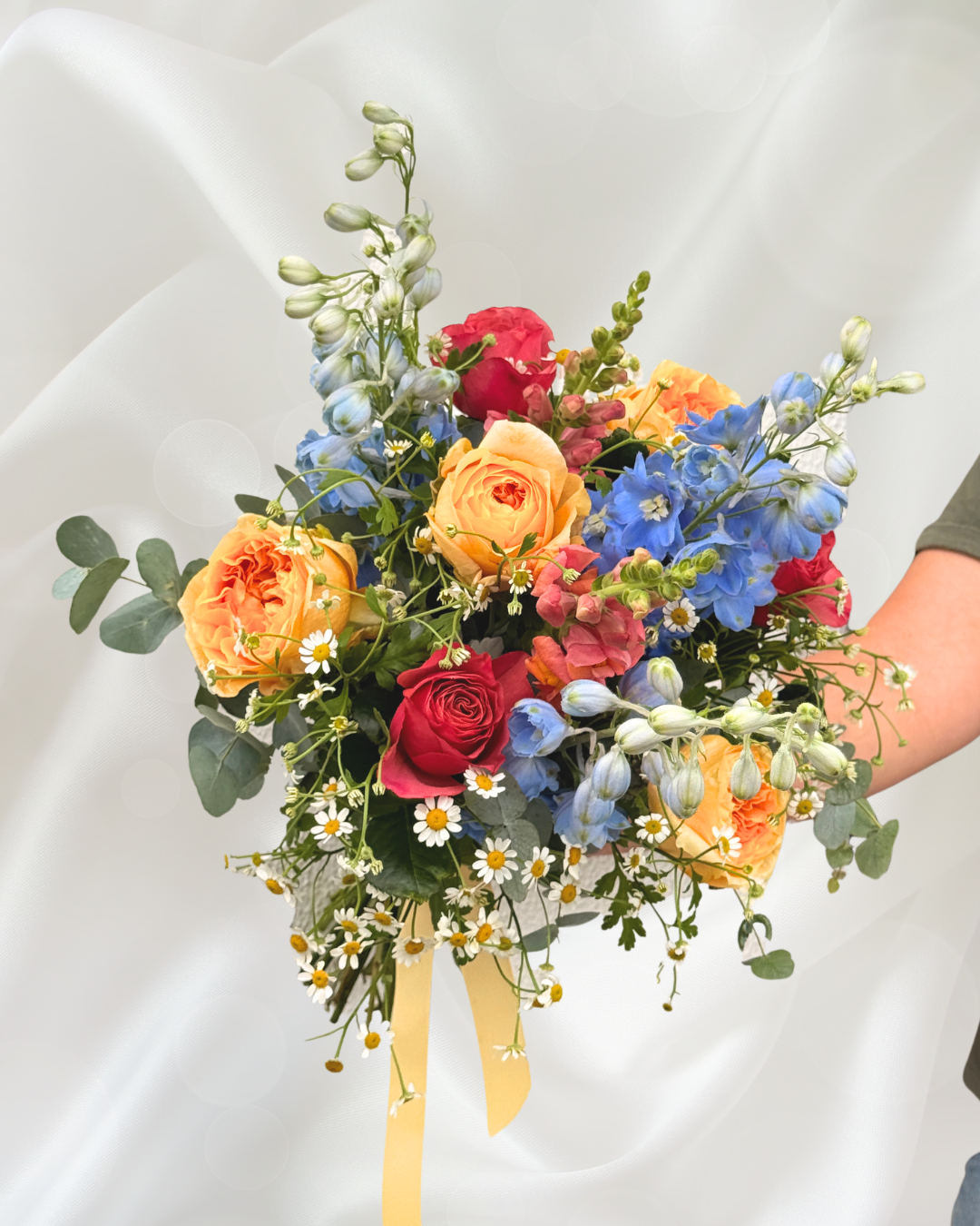 A colorful bouquet of flowers with roses, daisies, and blue delphiniums, held by a person, against a soft, white background.