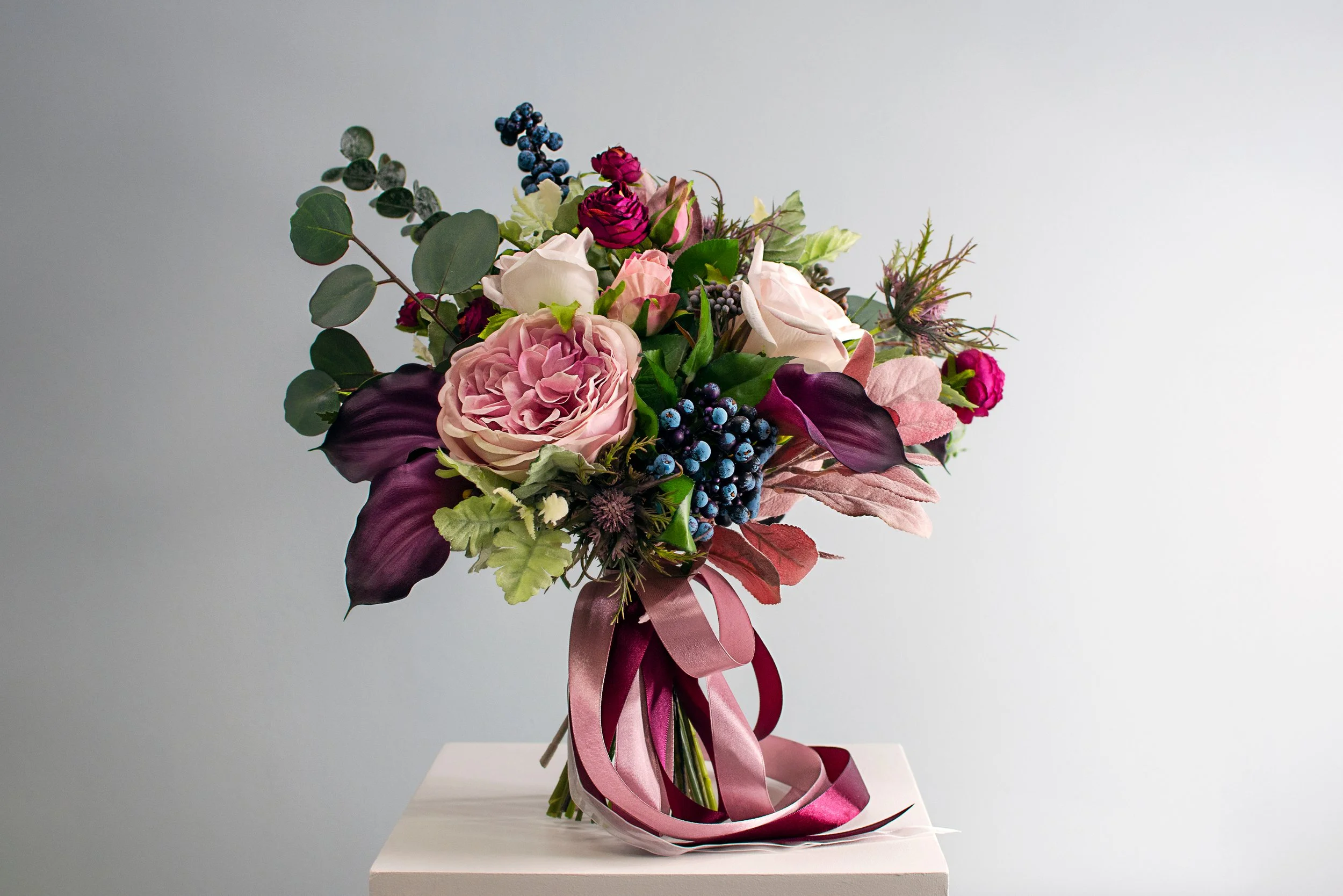 A bouquet of pink, purple, white, and green flowers with dark purple and green leaves, tied with pink and purple ribbons, placed on a white pedestal against a light gray background.