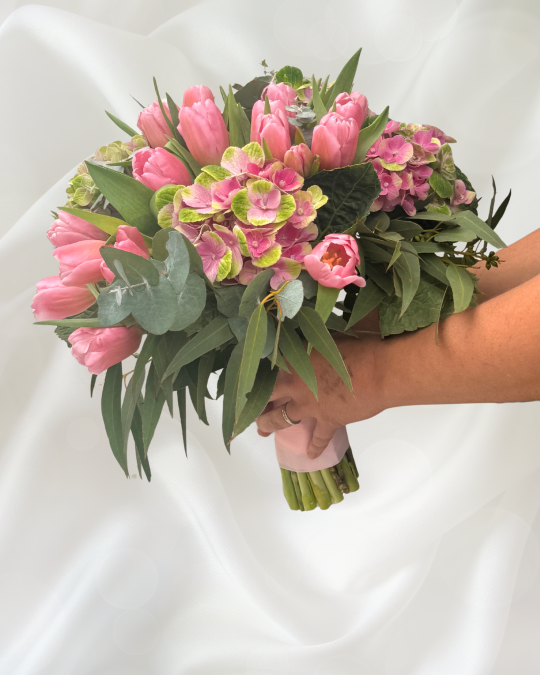 Person holding a bouquet of pink tulips, pink hydrangeas, and green foliage against a white background.