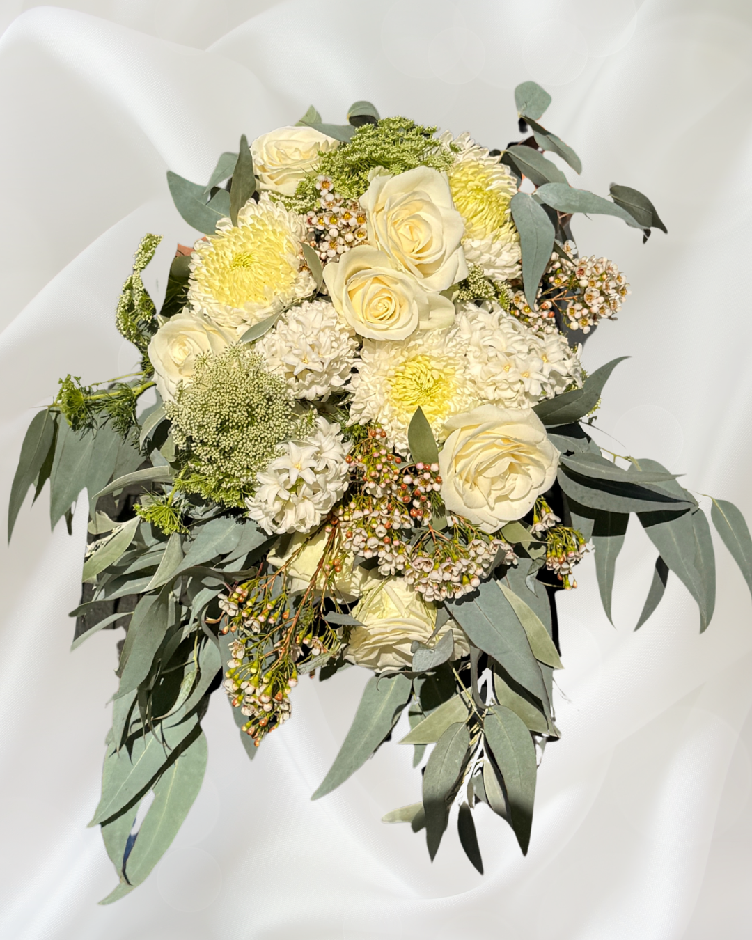 A floral arrangement with white roses, cream-colored roses, white hydrangeas, green foliage, and small pink and white berries, set against a white, satin-like background.