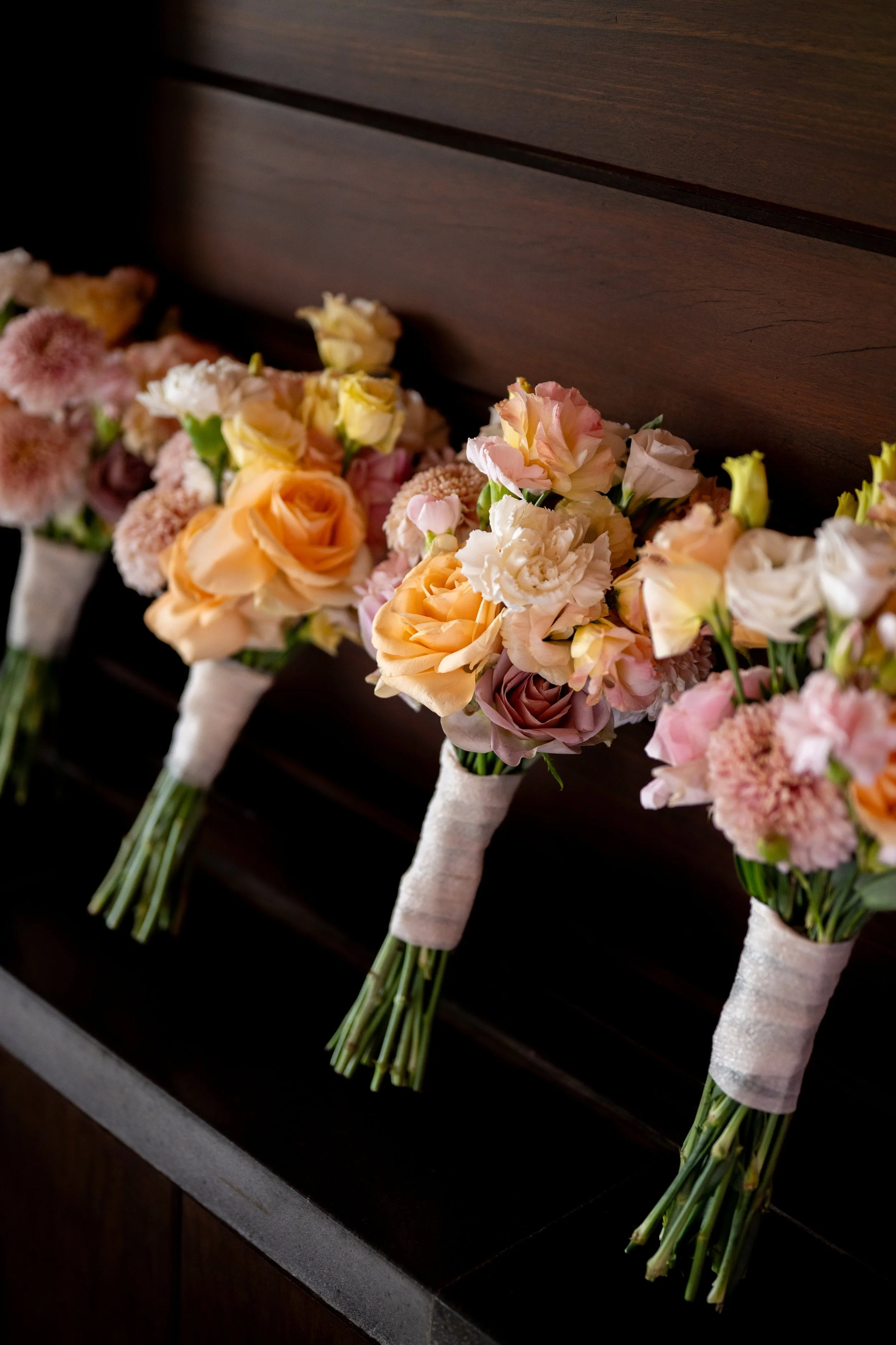 Five floral bouquets of pale pink, peach, and white flowers with green stems wrapped in white ribbon lie on a dark wooden surface.