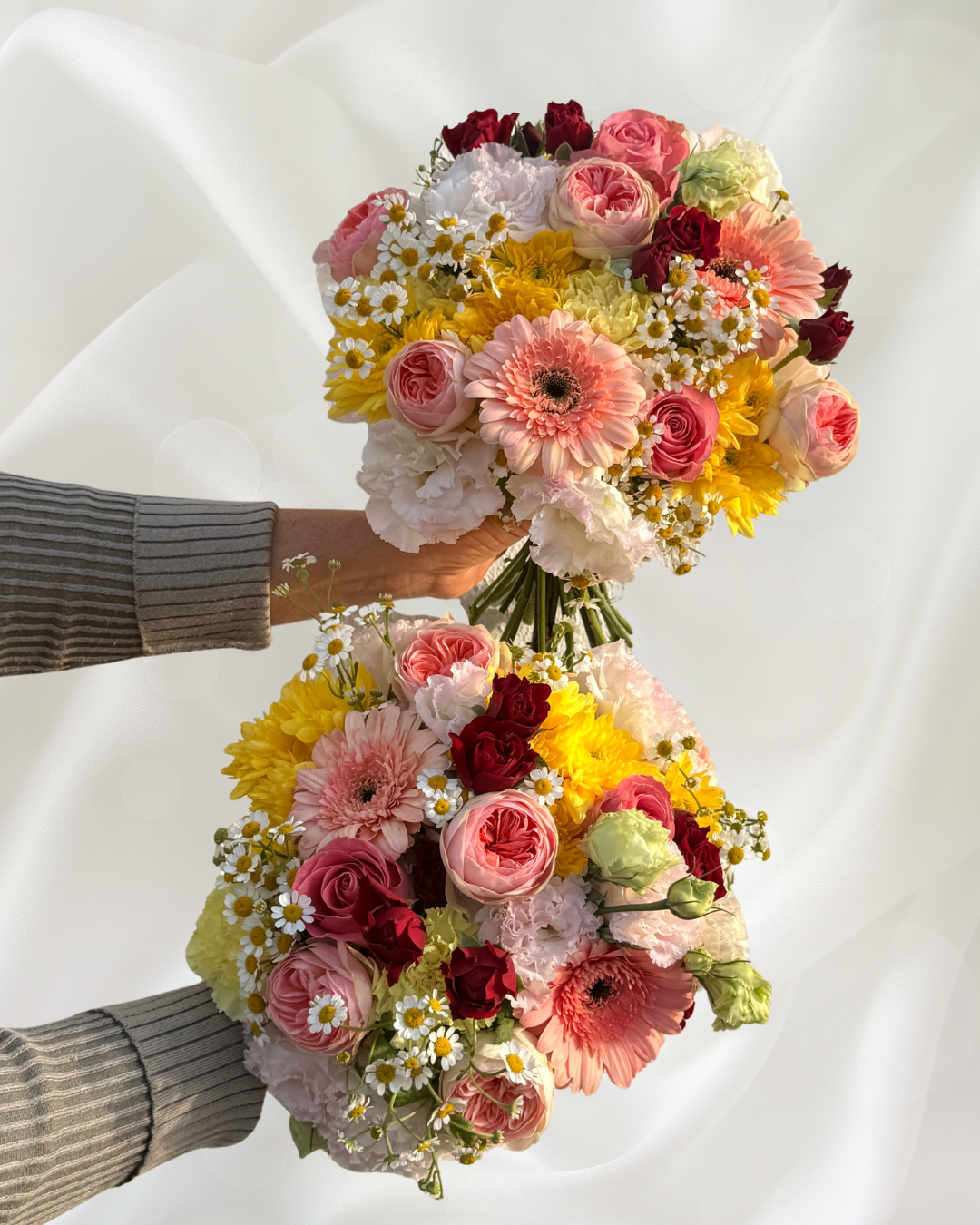 Two bouquets of colorful flowers, including roses, daisies, and gerbera daisies, held by two hands against a white background.