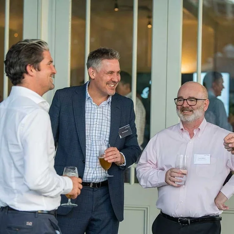 Three men in business attire are standing together, smiling, and holding drinks at a social event.