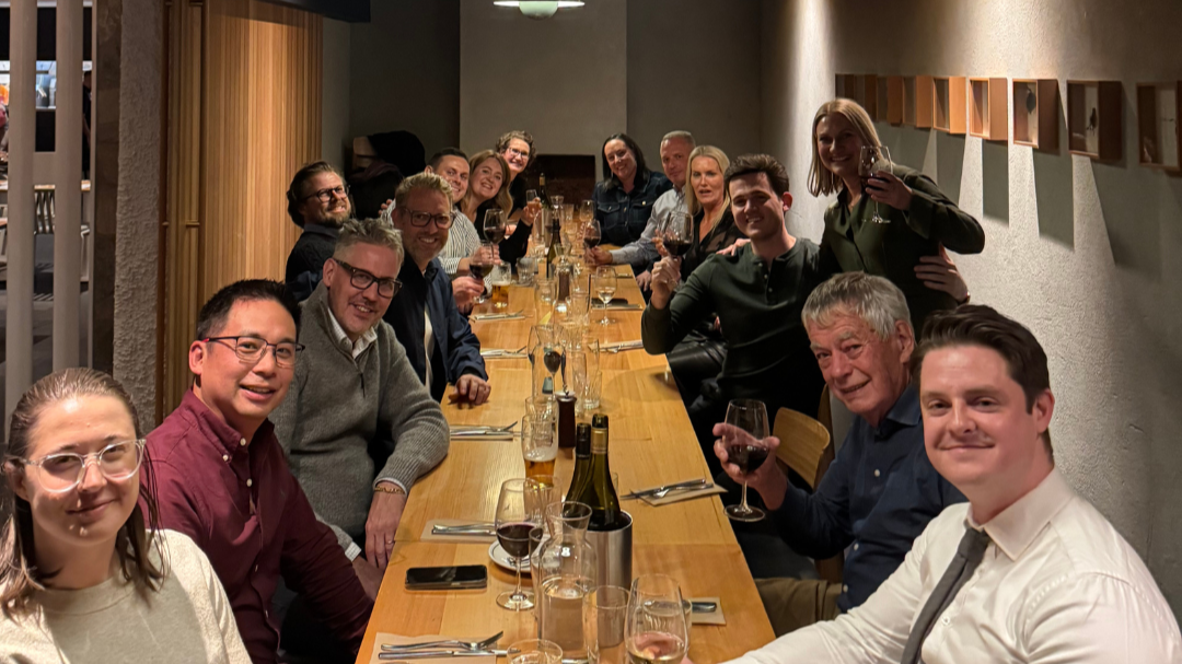 Group of people enjoying a dinner party at a long wooden table in a modern restaurant, holding wine glasses and smiling.