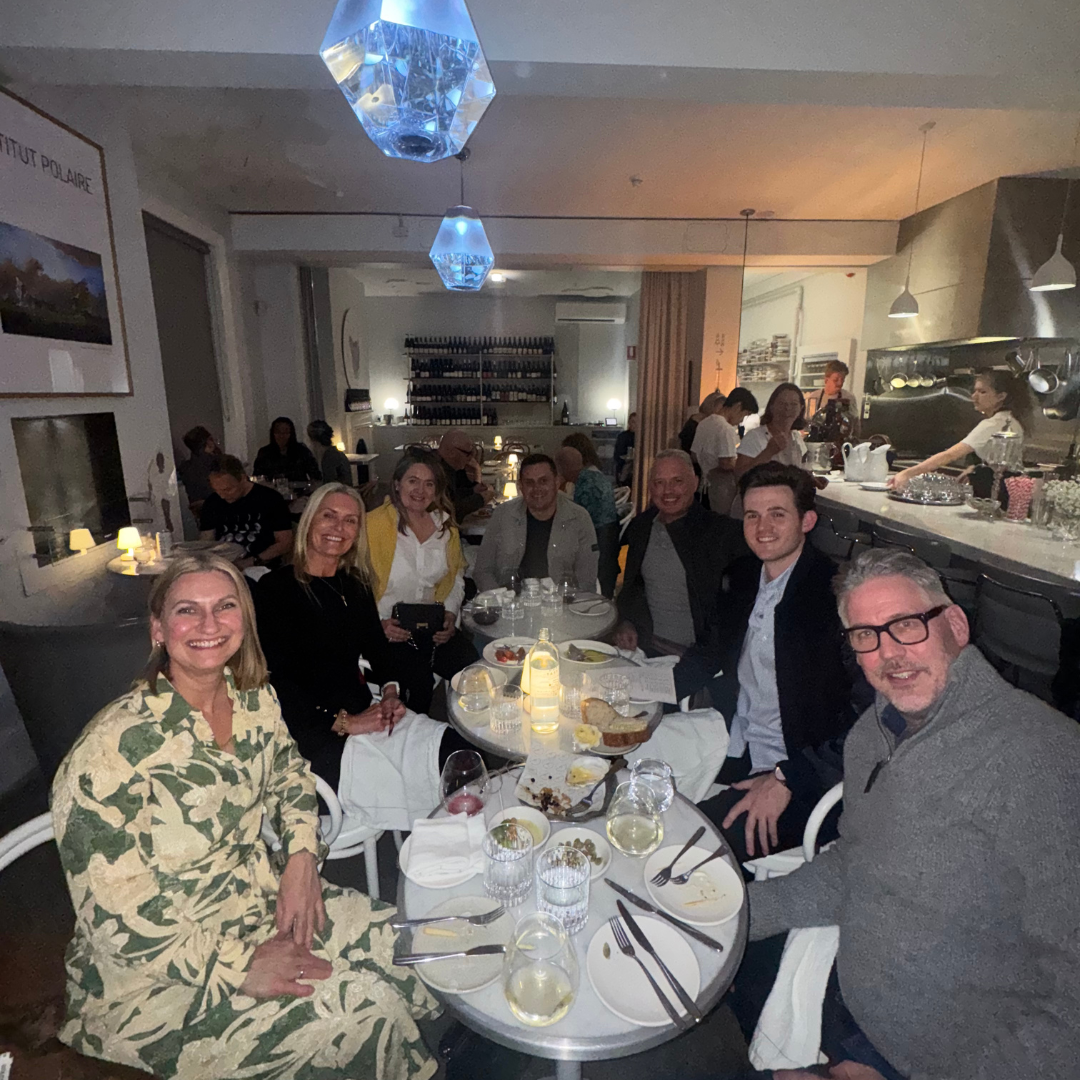A group of people sitting around a dining table in a restaurant at night, smiling for the camera, with food and drinks on the table, and a kitchen area with chefs in the background.