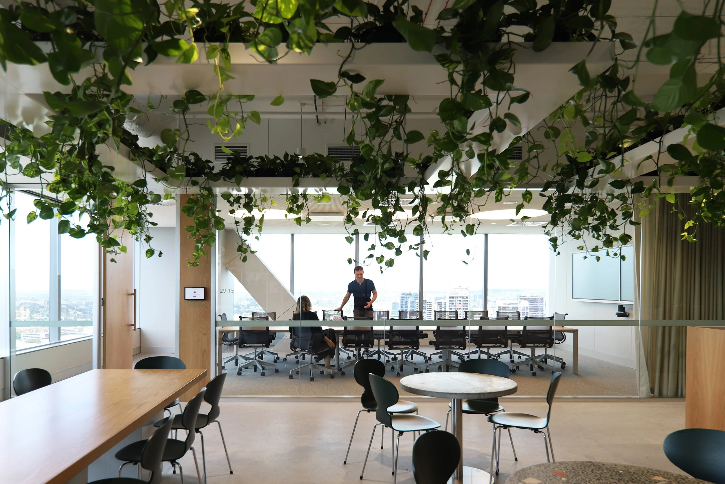 Modern office meeting room with large windows, a long table surrounded by chairs, and a man and woman engaged in conversation. Green plants hanging from the ceiling and a view of a city skyline outside.