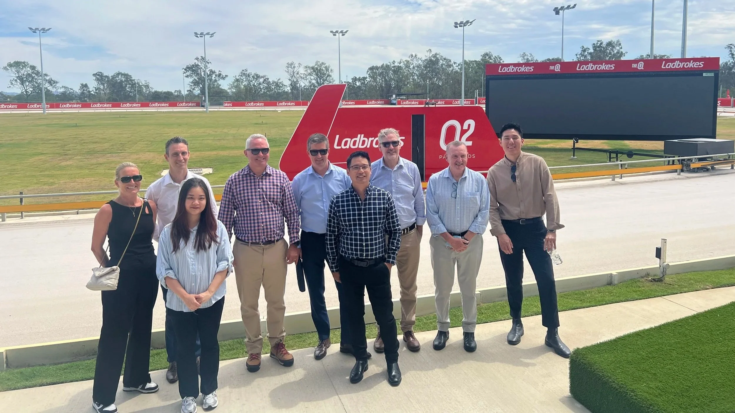Group of nine people standing in front of a race track with a large electronic scoreboard and signage for Ladbrokes, under a partly cloudy sky.