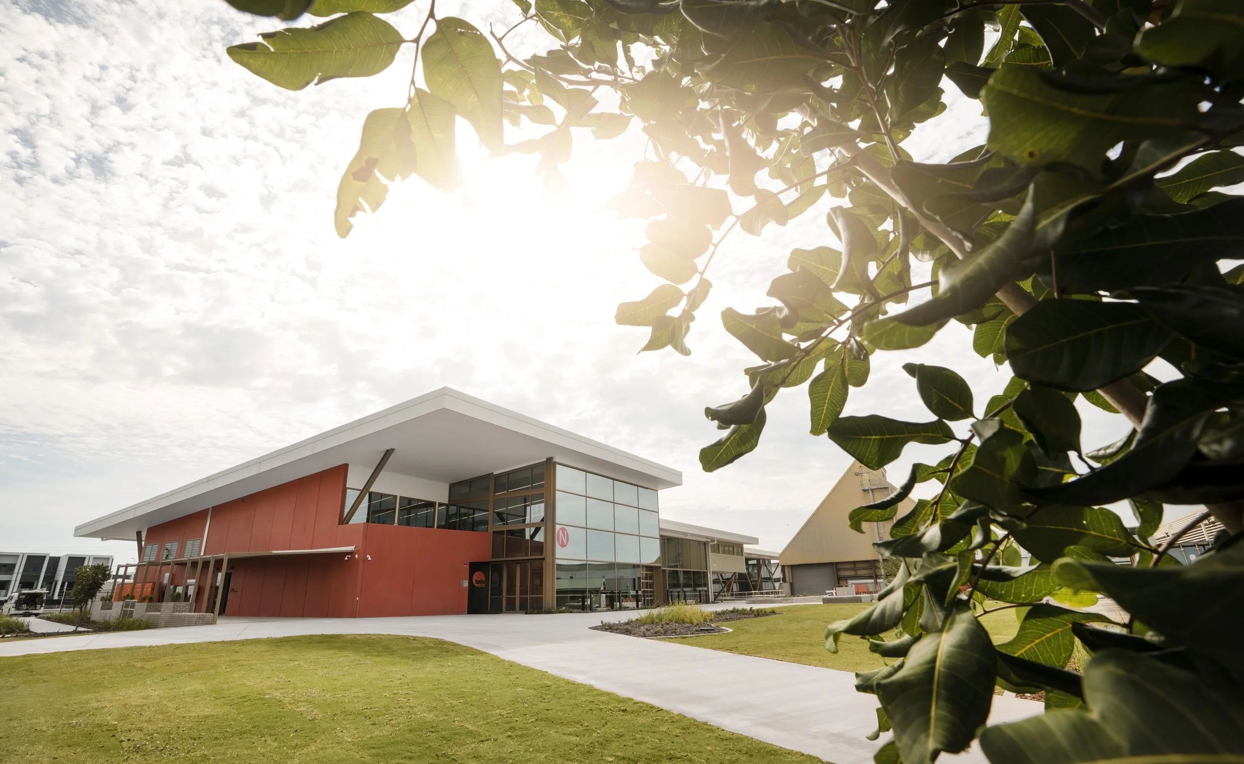 Modern building with large glass windows and red exterior walls, surrounded by a well-maintained lawn and tree in the foreground, with partly cloudy sky.