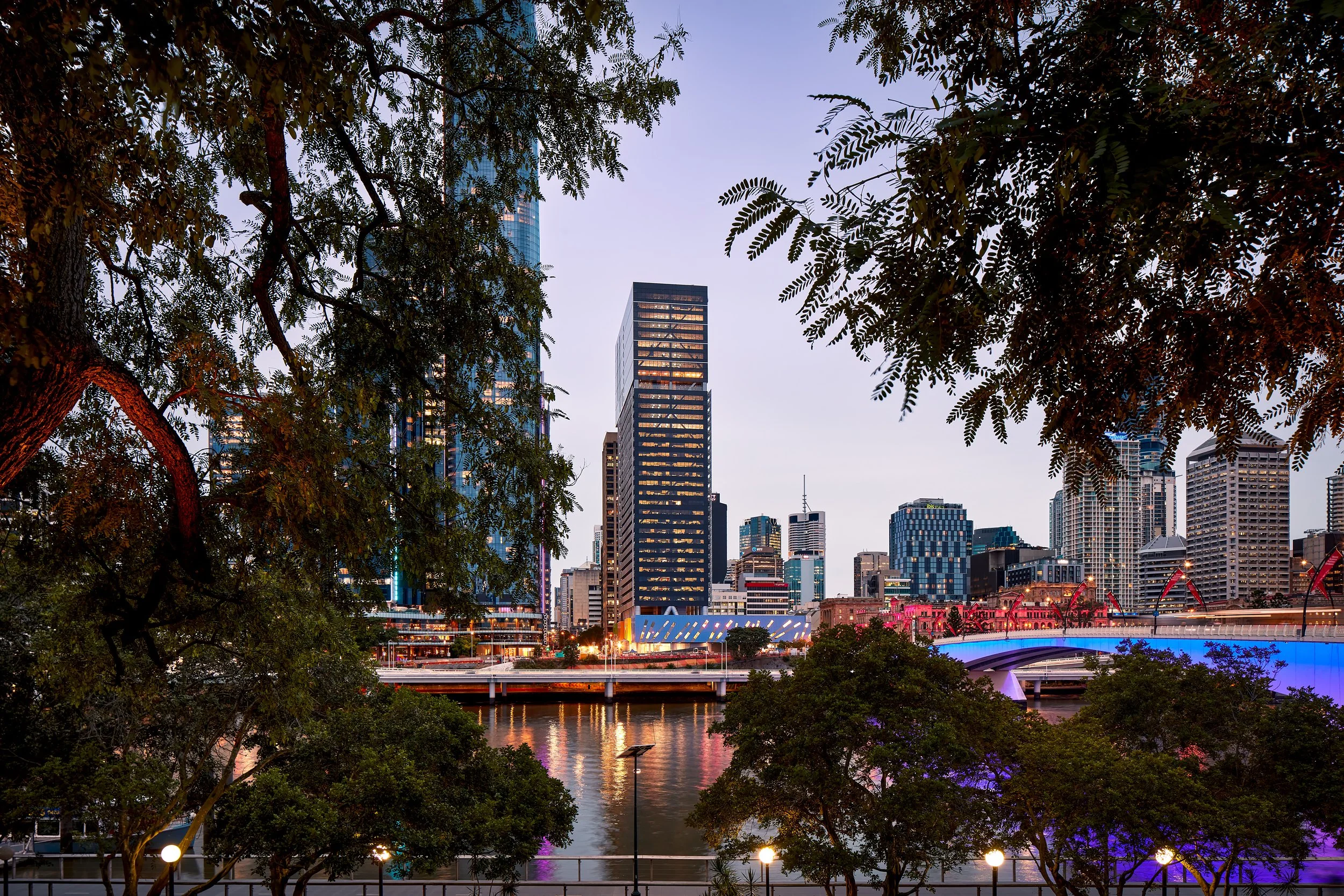 City skyline at dusk with tall buildings, a river in the foreground, and trees framing the scene.
