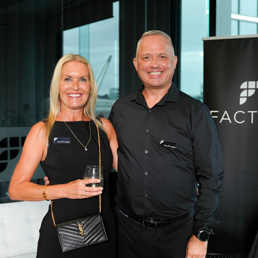 A man and woman smiling and posing together at an indoor event, with a window in the background. The woman is holding a glass of wine and wearing a black dress with a Yves Saint Laurent purse. The man is wearing a black shirt and has a name tag. They are standing close with their arms around each other.