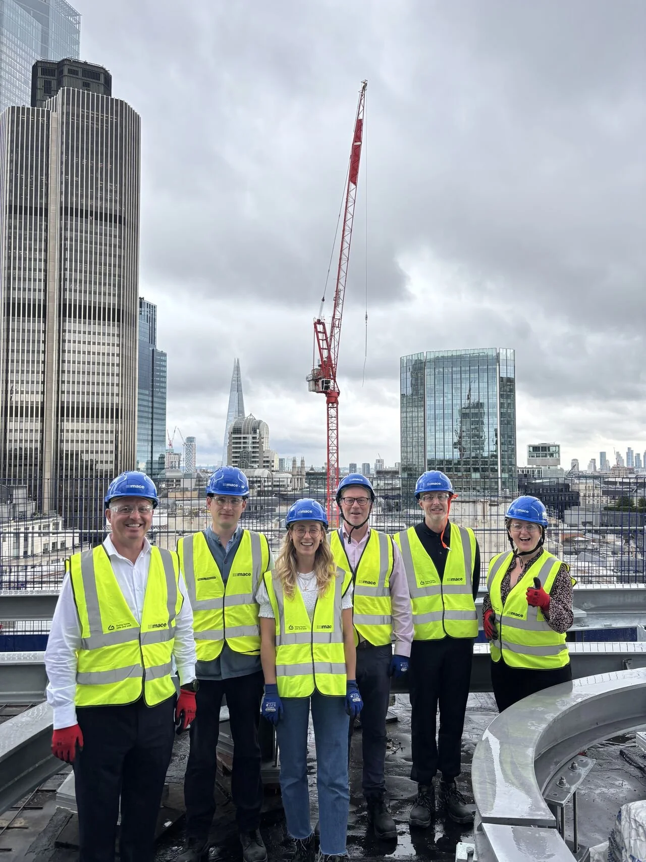 Group of six construction workers wearing yellow safety vests and blue hard hats, standing on a building rooftop with a cityscape of tall buildings, a crane, and cloudy sky in the background.