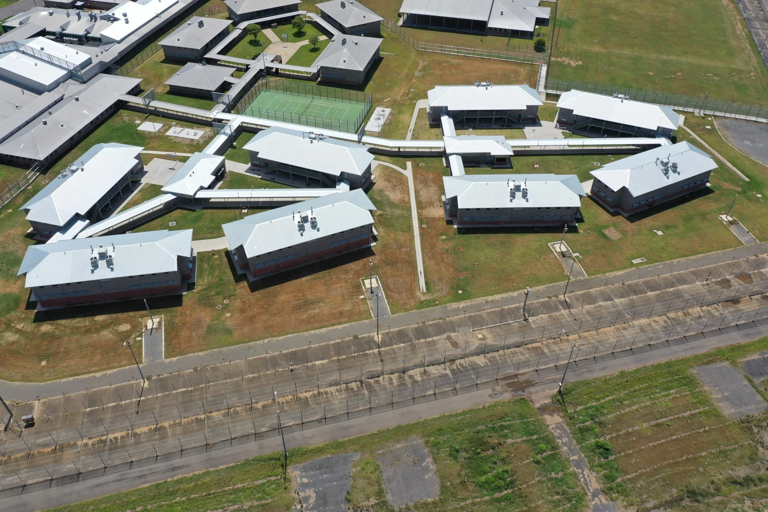 Aerial view of a prison complex with multiple buildings, a tennis court, a fenced perimeter, and surrounding open land.