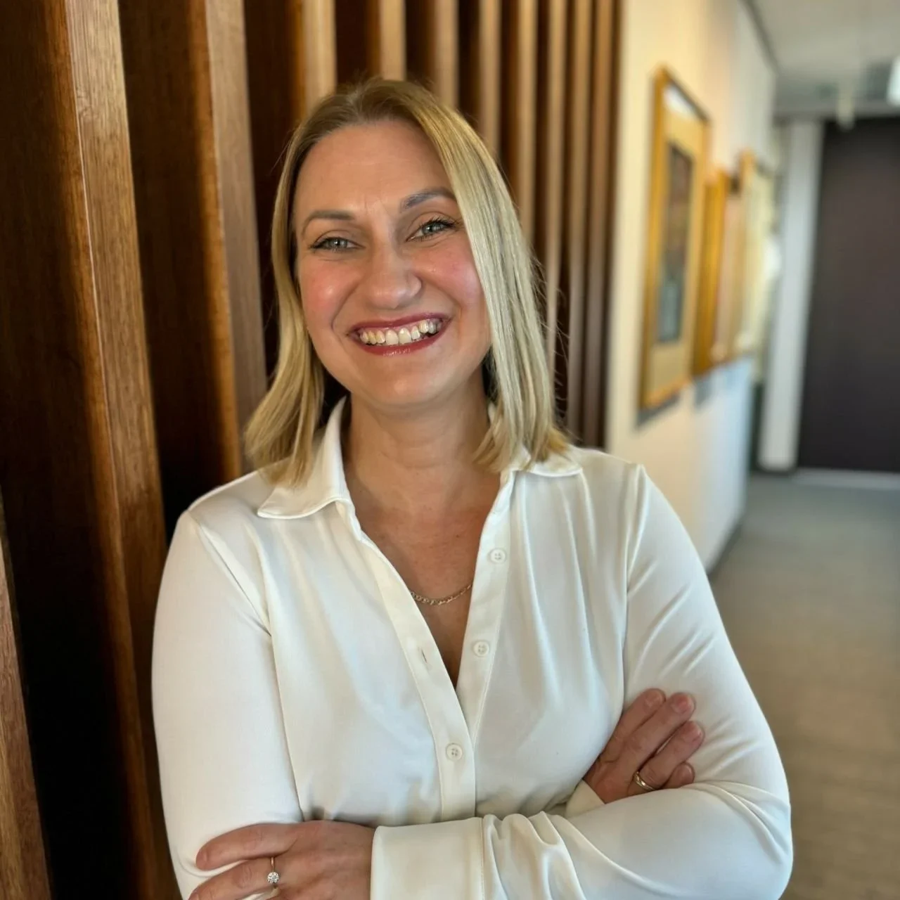 A woman with shoulder-length blonde hair, wearing a white button-down shirt, standing in an indoor hallway with wooden wall panels and framed pictures.
