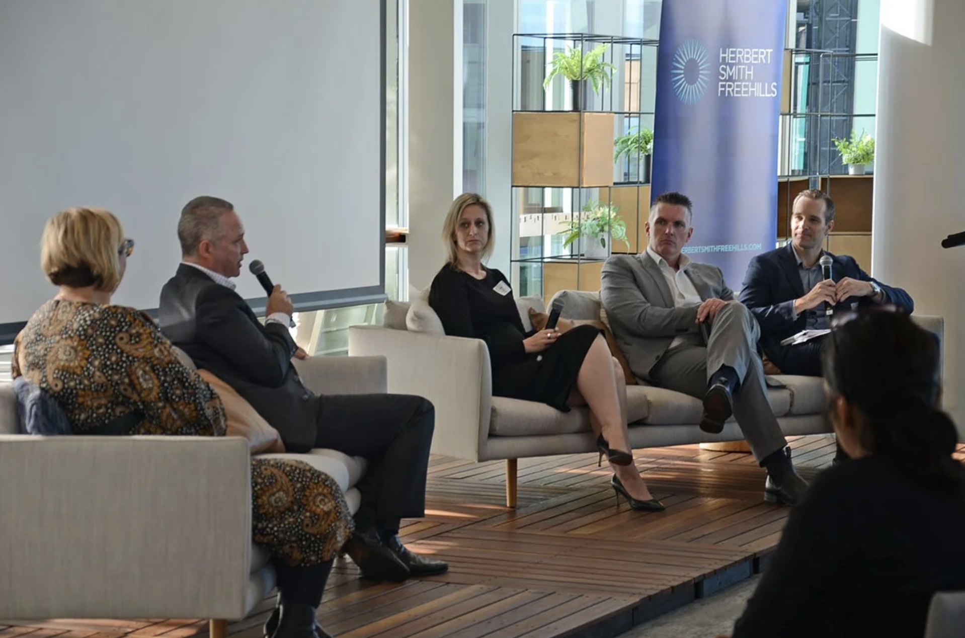 Panel discussion with five individuals seated on a stage in front of a large window, with a banner reading 'Herbert Smith Freehills' in the background.