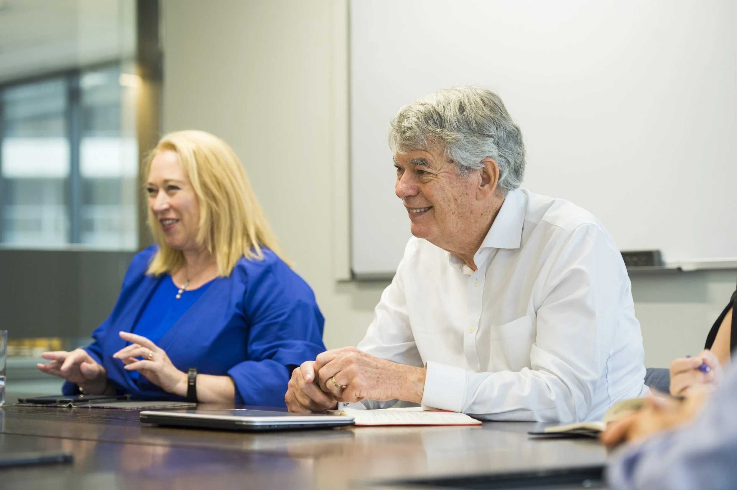 Two people sitting at a conference table in a meeting room, smiling and engaged in a discussion. The woman on the left has blonde hair and is wearing a blue top, while the man on the right has gray hair and is dressed in a white shirt.