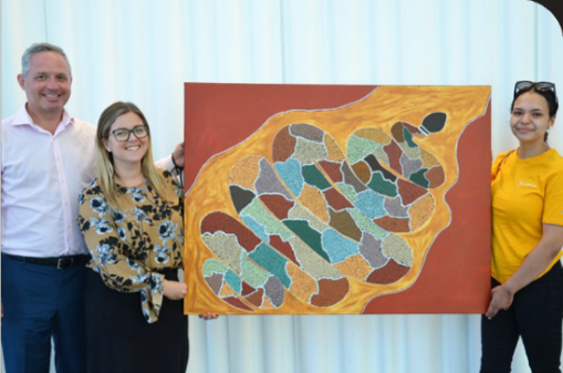 Three people standing outdoors next to a colorful abstract painting on a red background.