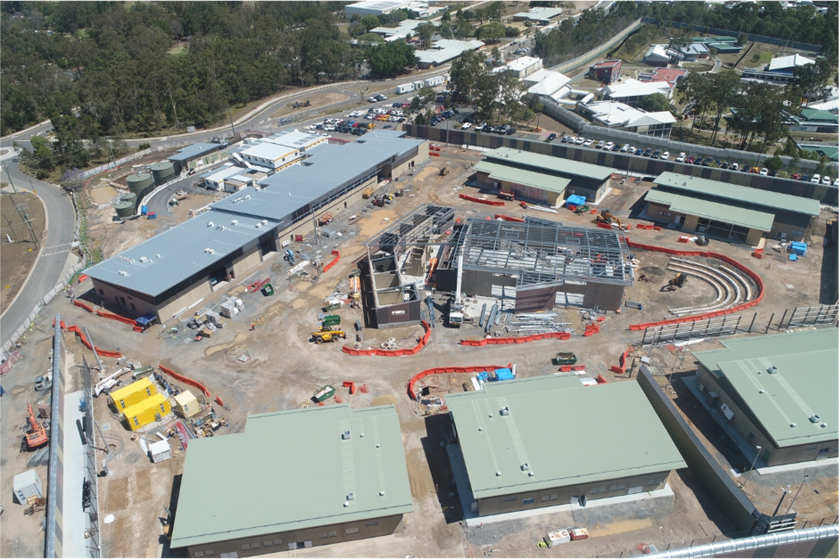 Aerial view of a construction site with several buildings in various stages of construction, surrounded by dirt, construction equipment, and orange safety barriers, with completed buildings and parking lot in the background.