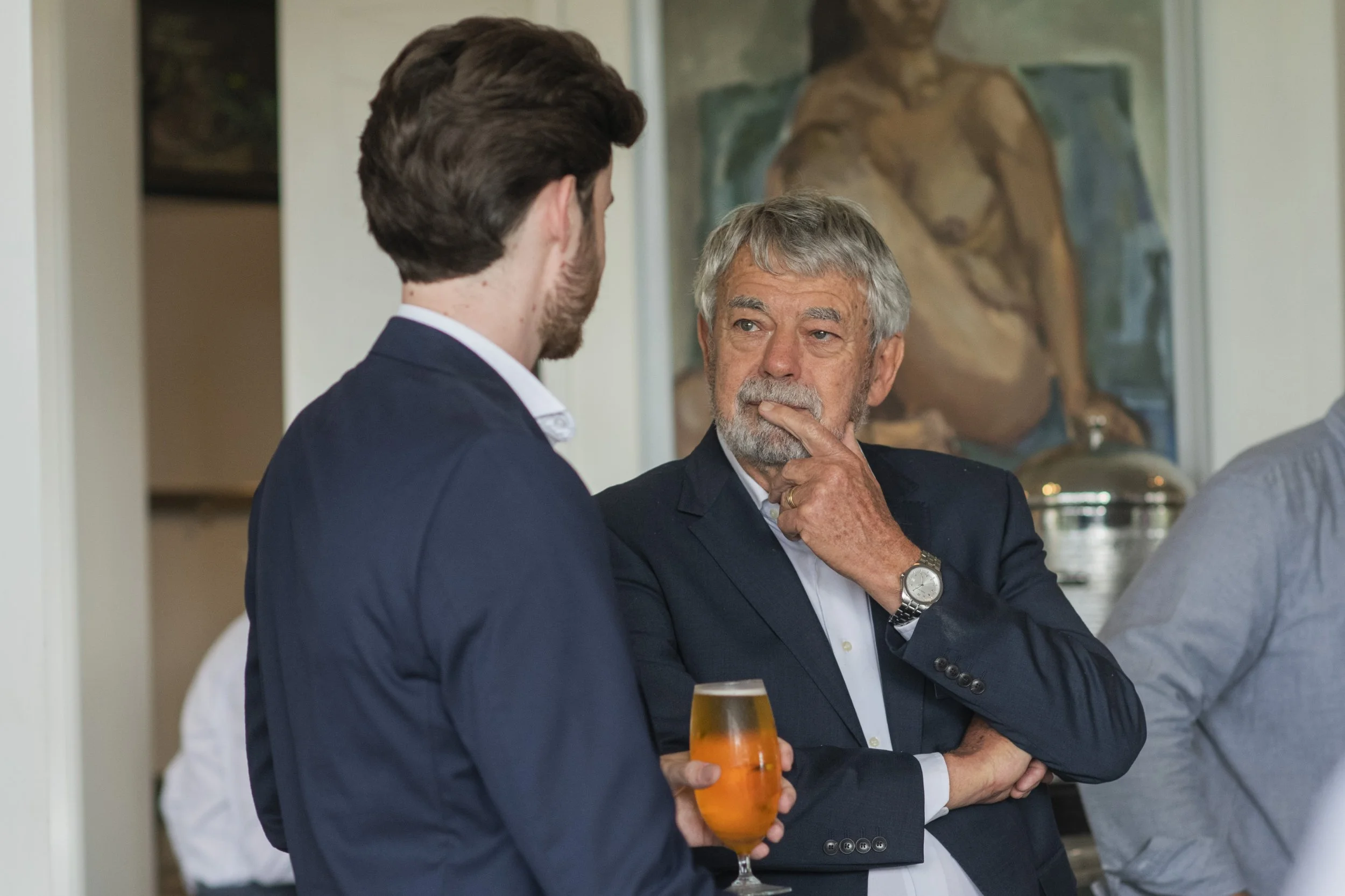Two men in suits engaged in conversation at a social gathering, one holding a glass of orange-colored drink, with a portrait painting in the background.