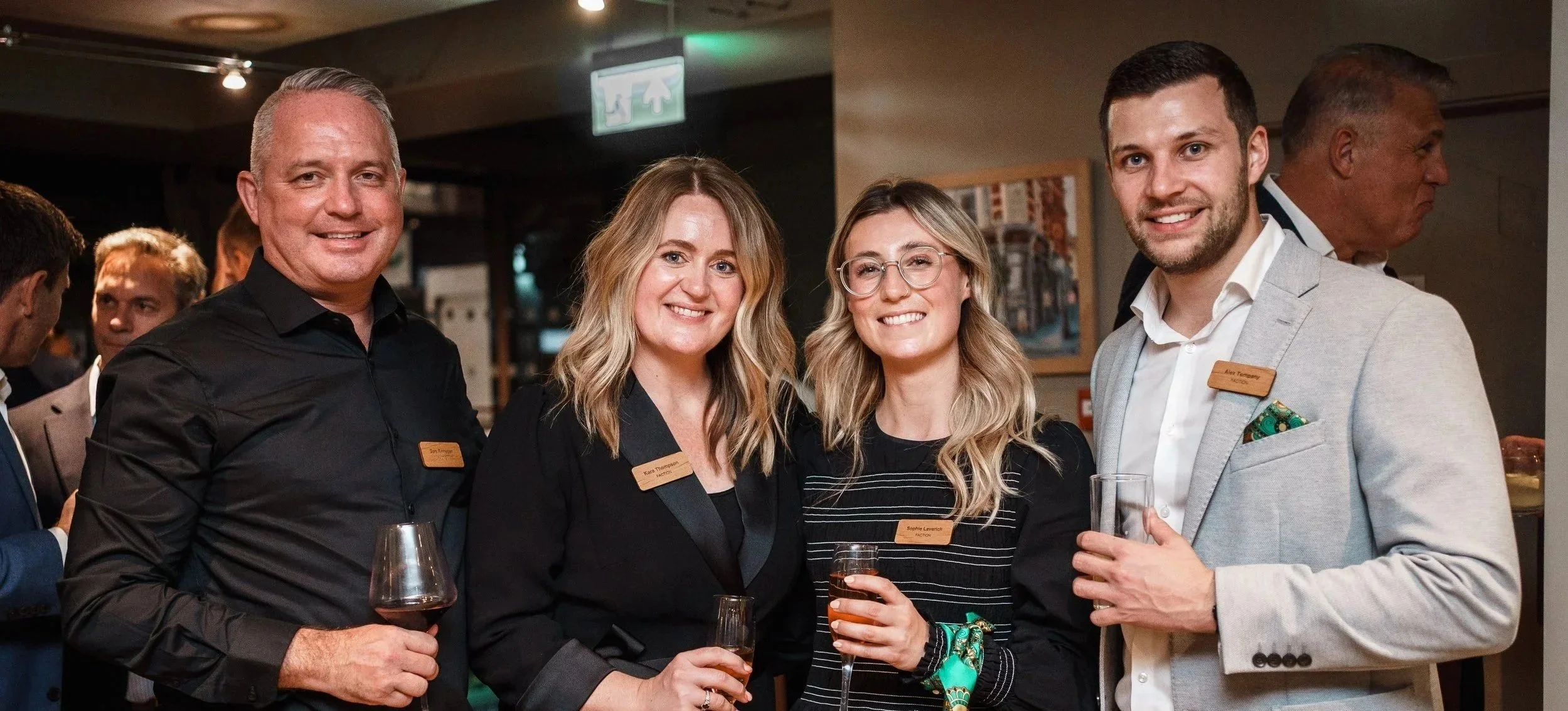 Group of four people at a social gathering, standing close together, holding drinks, smiling for the photo, in an indoor venue.