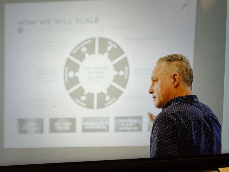 A man with short blond hair, wearing a dark polka dot shirt, pointing at a projected presentation slide on a wall, which features a circular diagram and text.