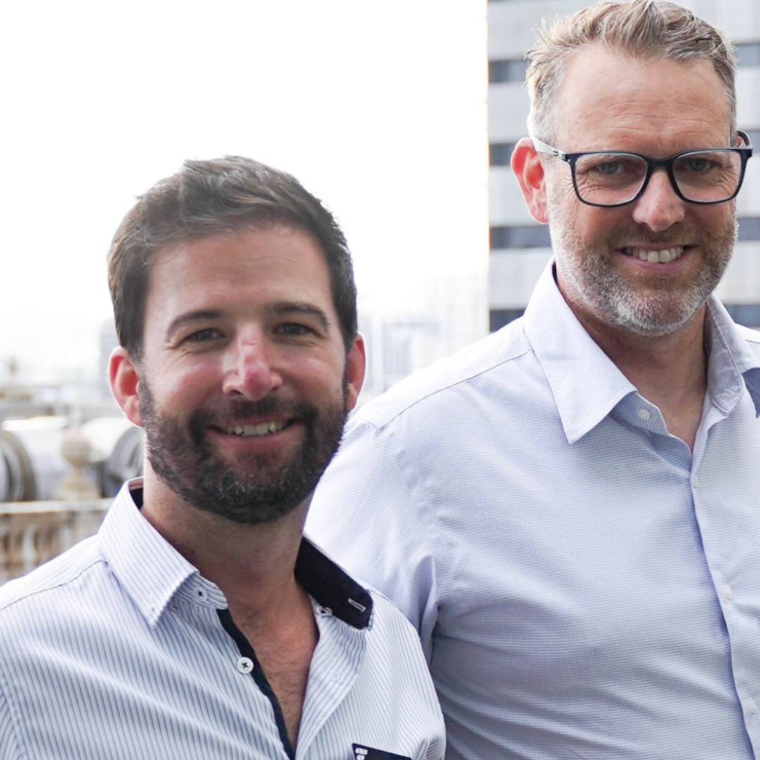 Two smiling men in business casual attire standing outdoors in an urban setting.