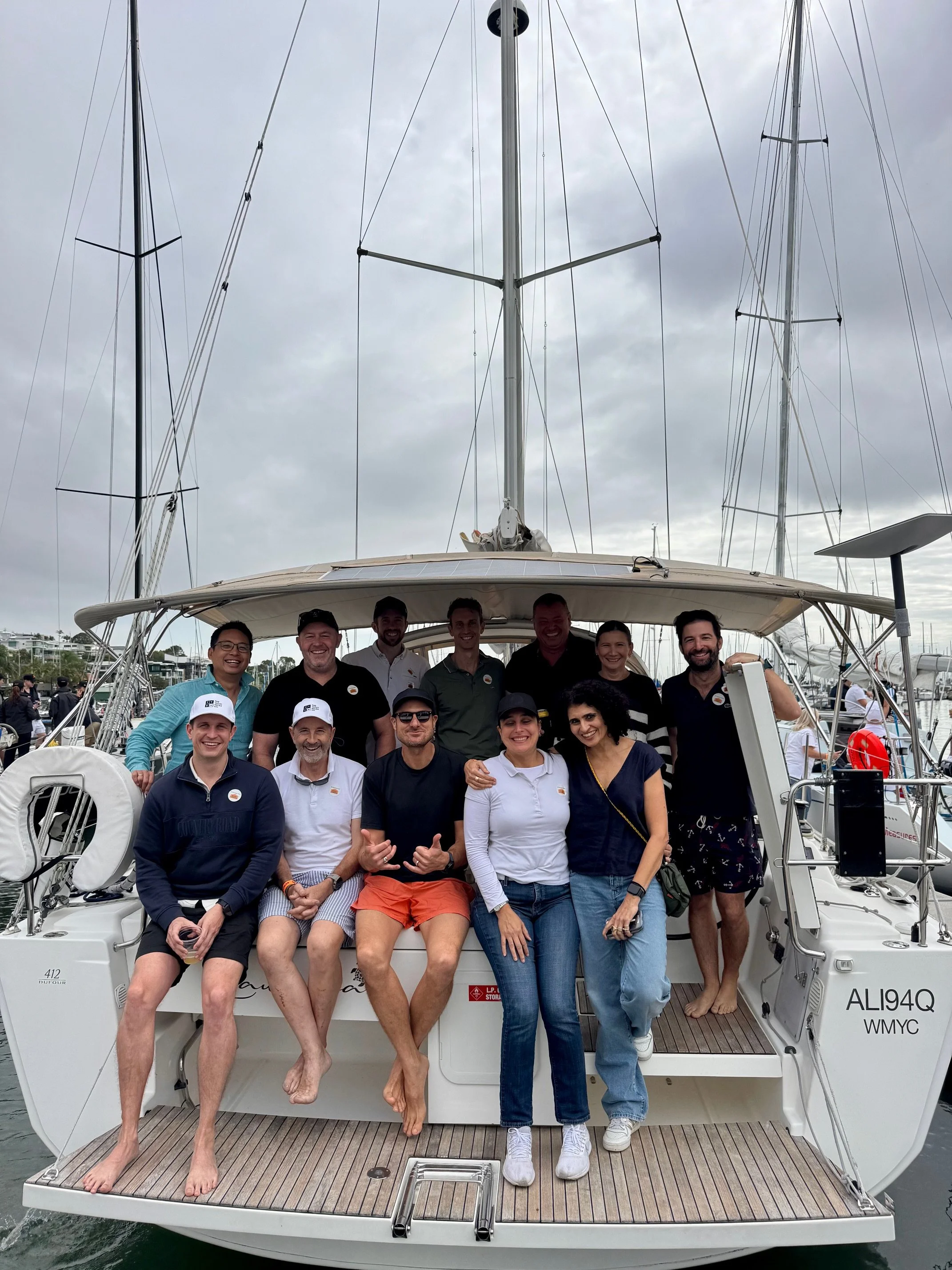 Group of people standing and sitting on the back of a sailboat docked at a marina, with cloudy sky overhead.