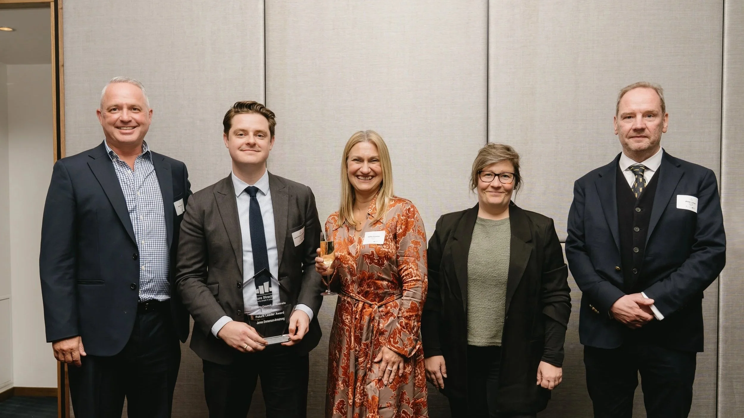 Group of five professionally dressed people standing together at an event, with one holding a glass of champagne and an award.