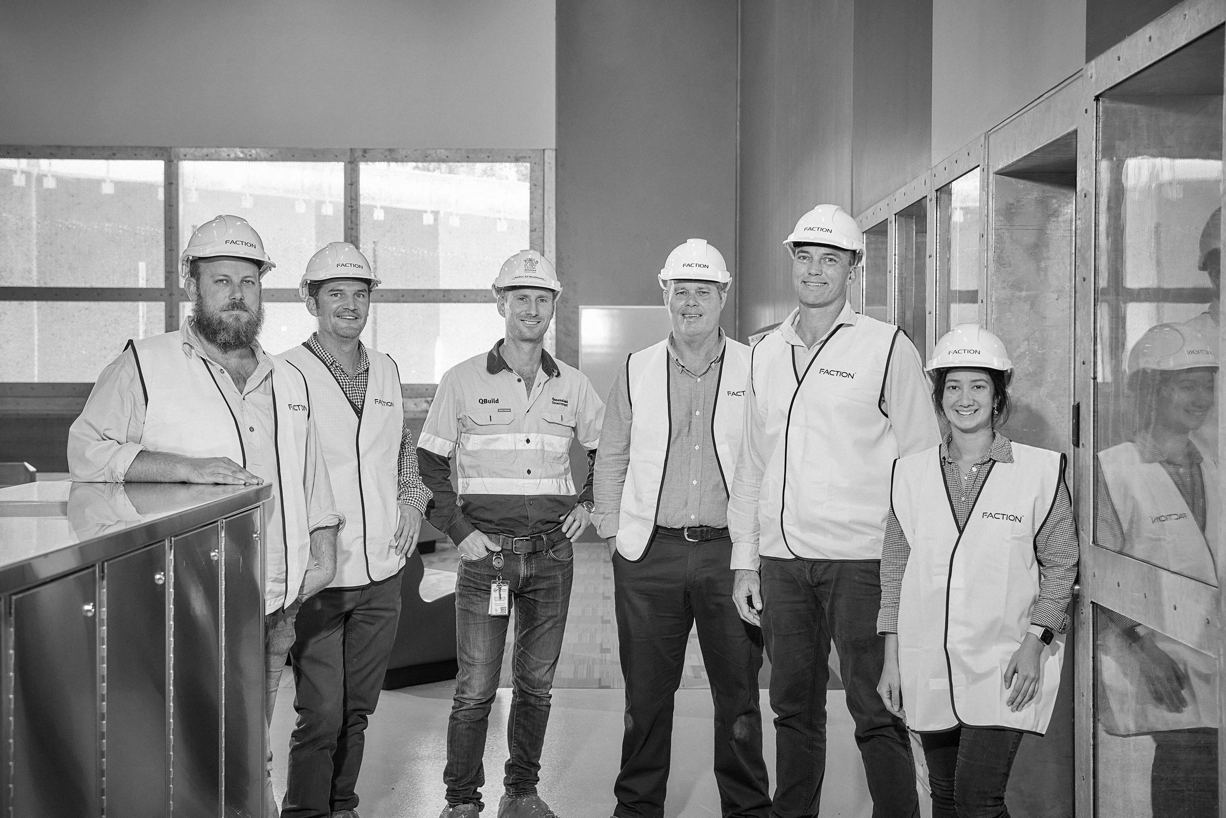 Group of seven construction workers standing inside a building, wearing safety helmets and vests, smiling at the camera.