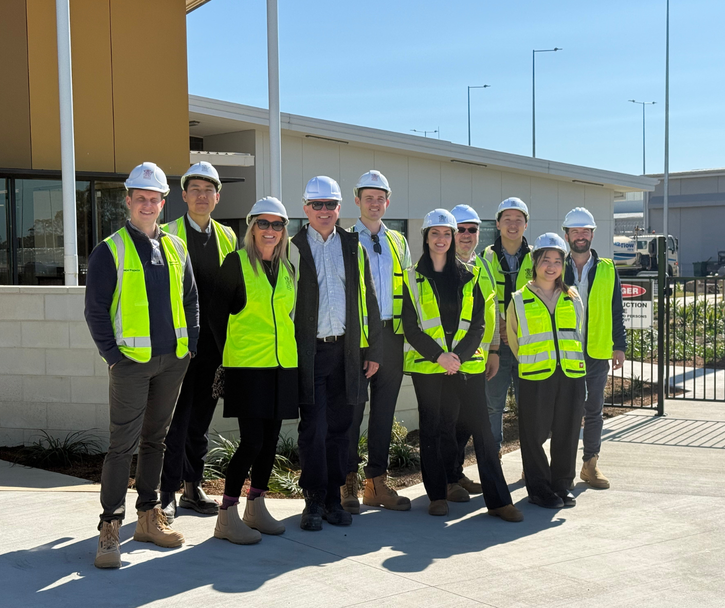 Group of ten people wearing safety vests and hard hats standing outside building on a sunny day.