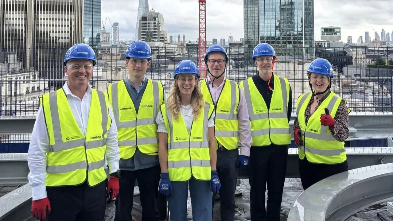 Six people wearing blue safety helmets, yellow high-visibility vests, and gloves on a construction site with a city skyline in the background, including the Salesforce Tower.