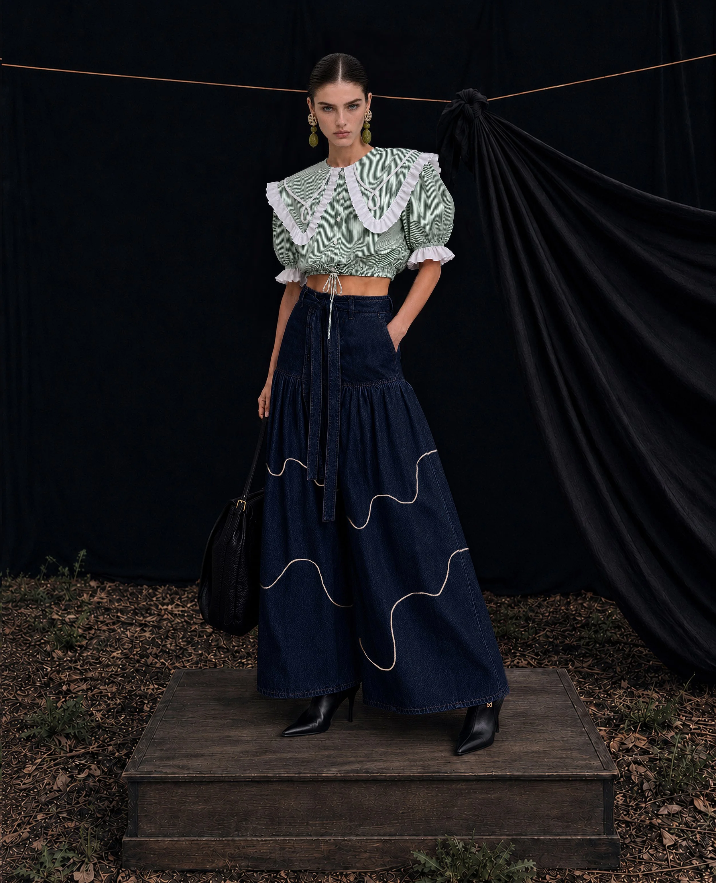 A female model standing outdoors on a wooden platform, wearing a green and white striped cropped blouse with puffed sleeves and ruffled white trim, dark high-waisted jeans, black high-heeled boots, large green dangle earrings, and carrying a black handbag, with black draped fabric behind her and a ground covered in leaves and small plants.